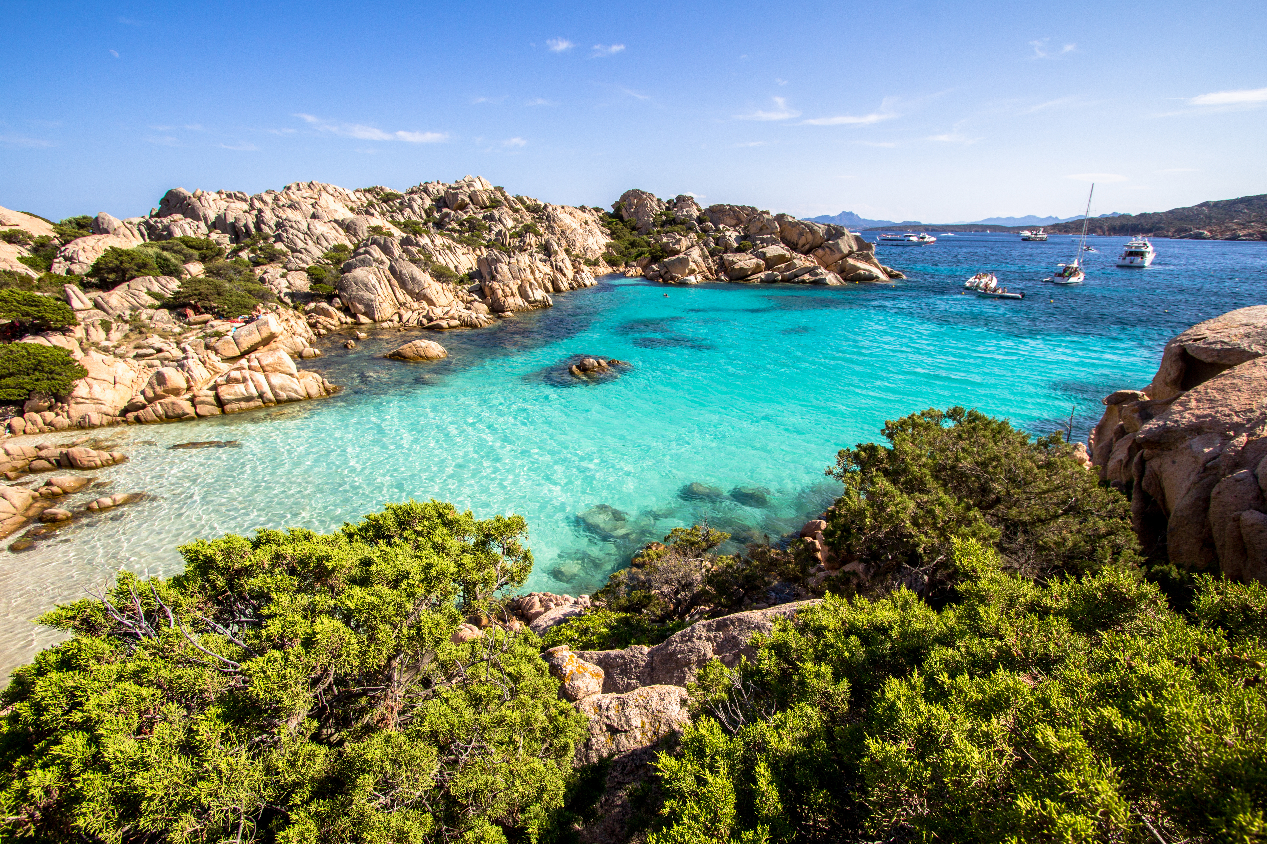 Clear turquoise bay, rocky shoreline, green shrubs, and distant boats.