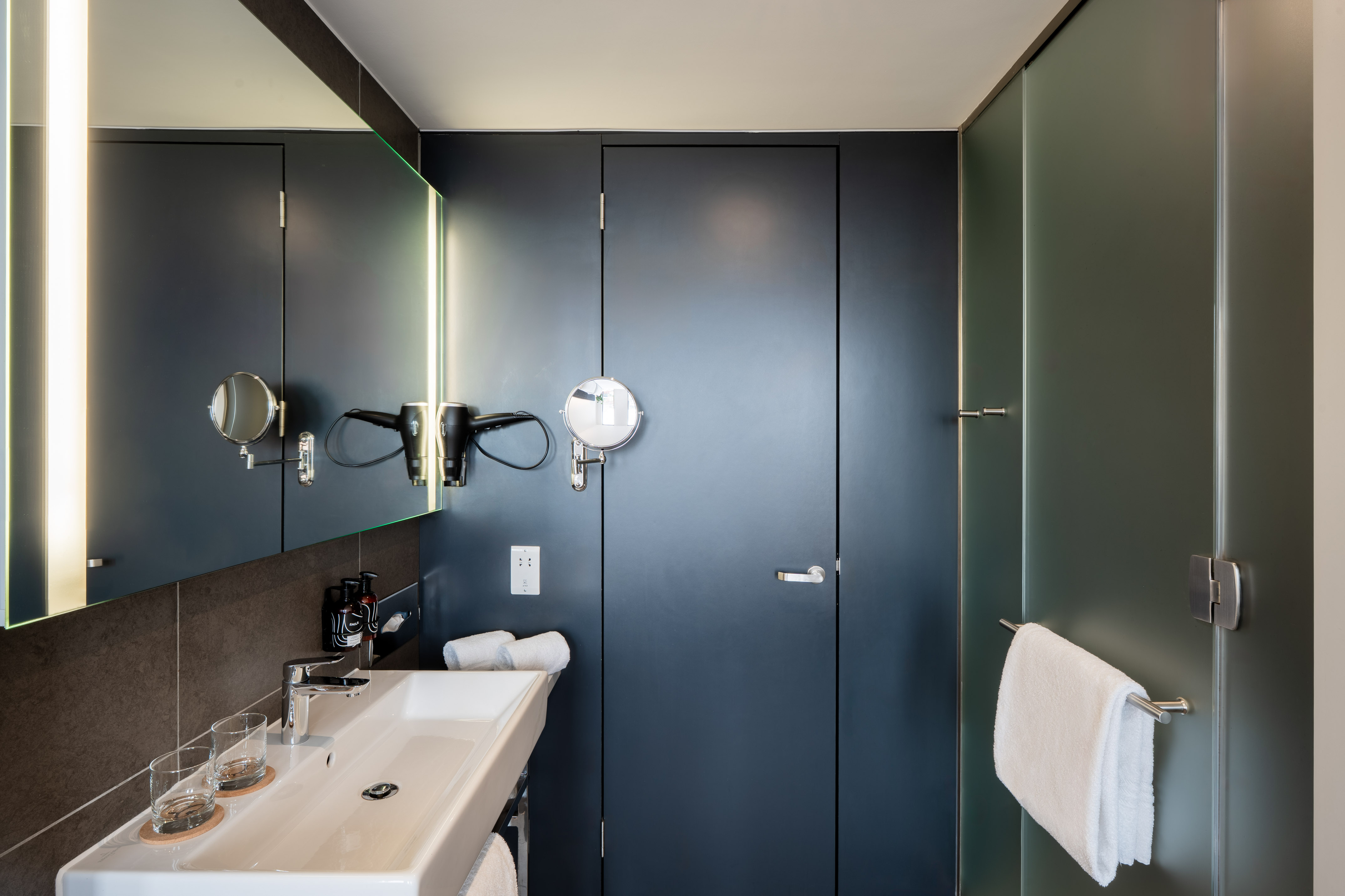 Modern bathroom with white sink, backlit mirror, dark decor, and glass shower.