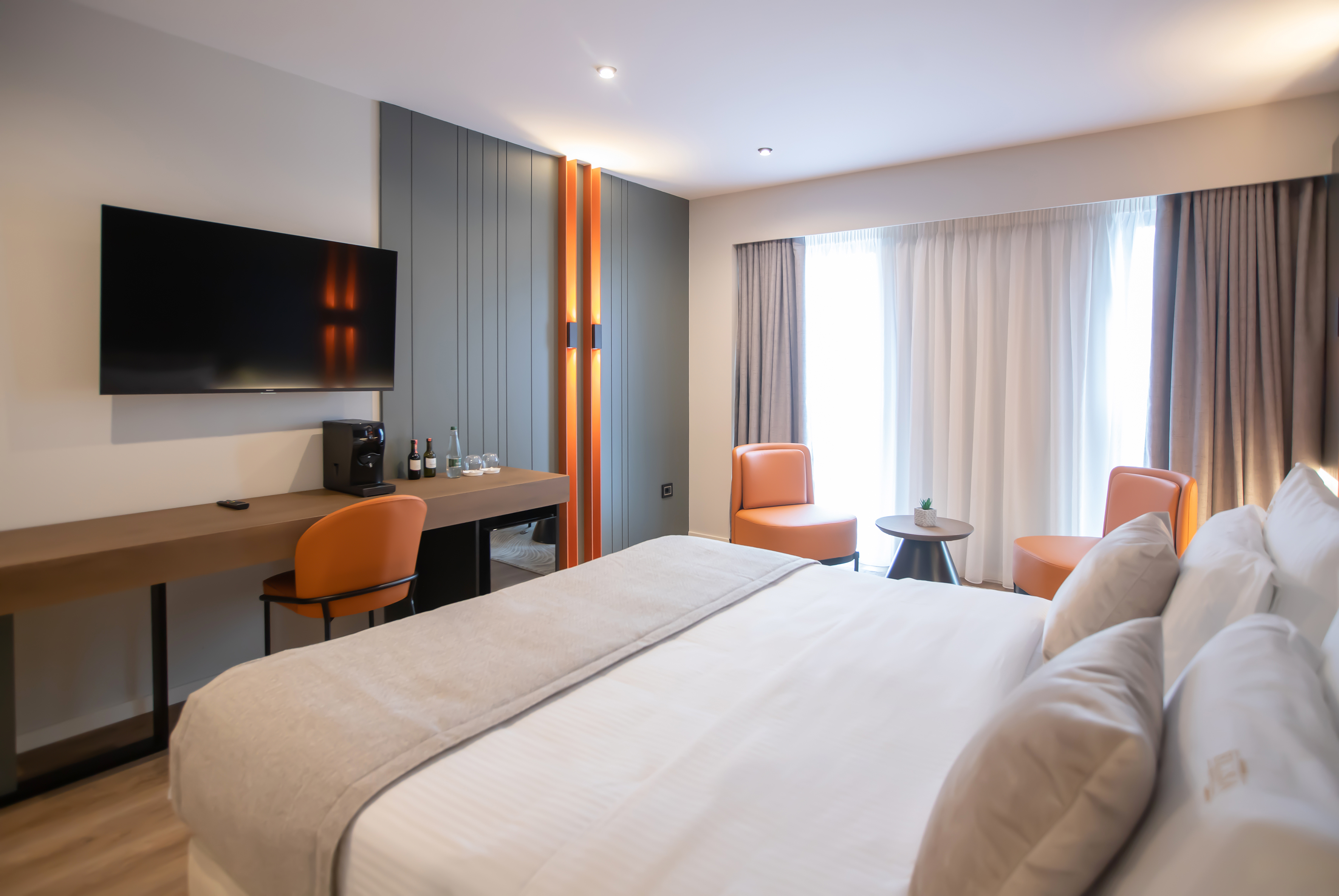 Modern hotel room with white bed, orange accent chairs, desk, and large TV.