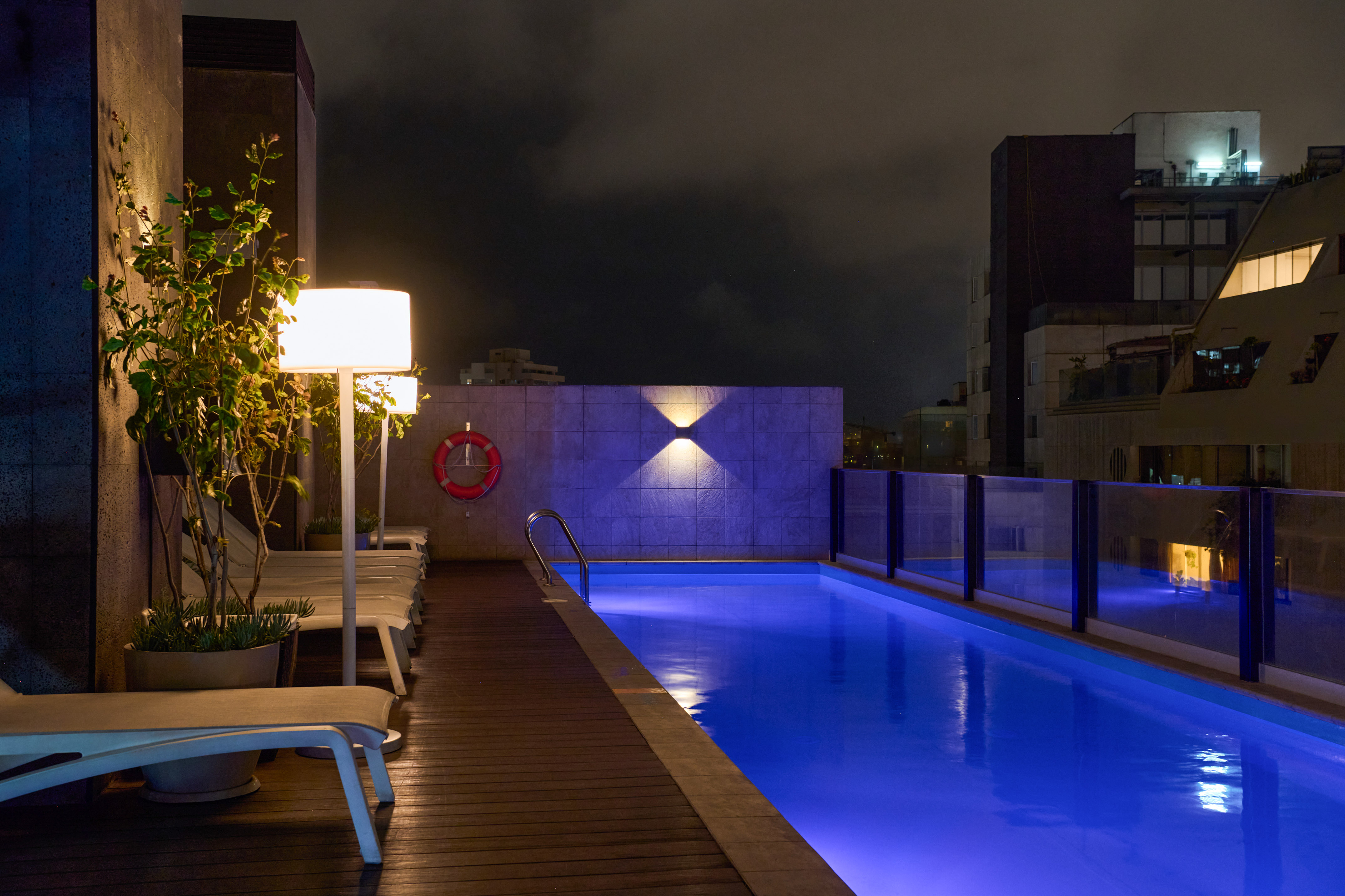 Illuminated rooftop pool at night with blue water, wooden deck, and lounge chairs.