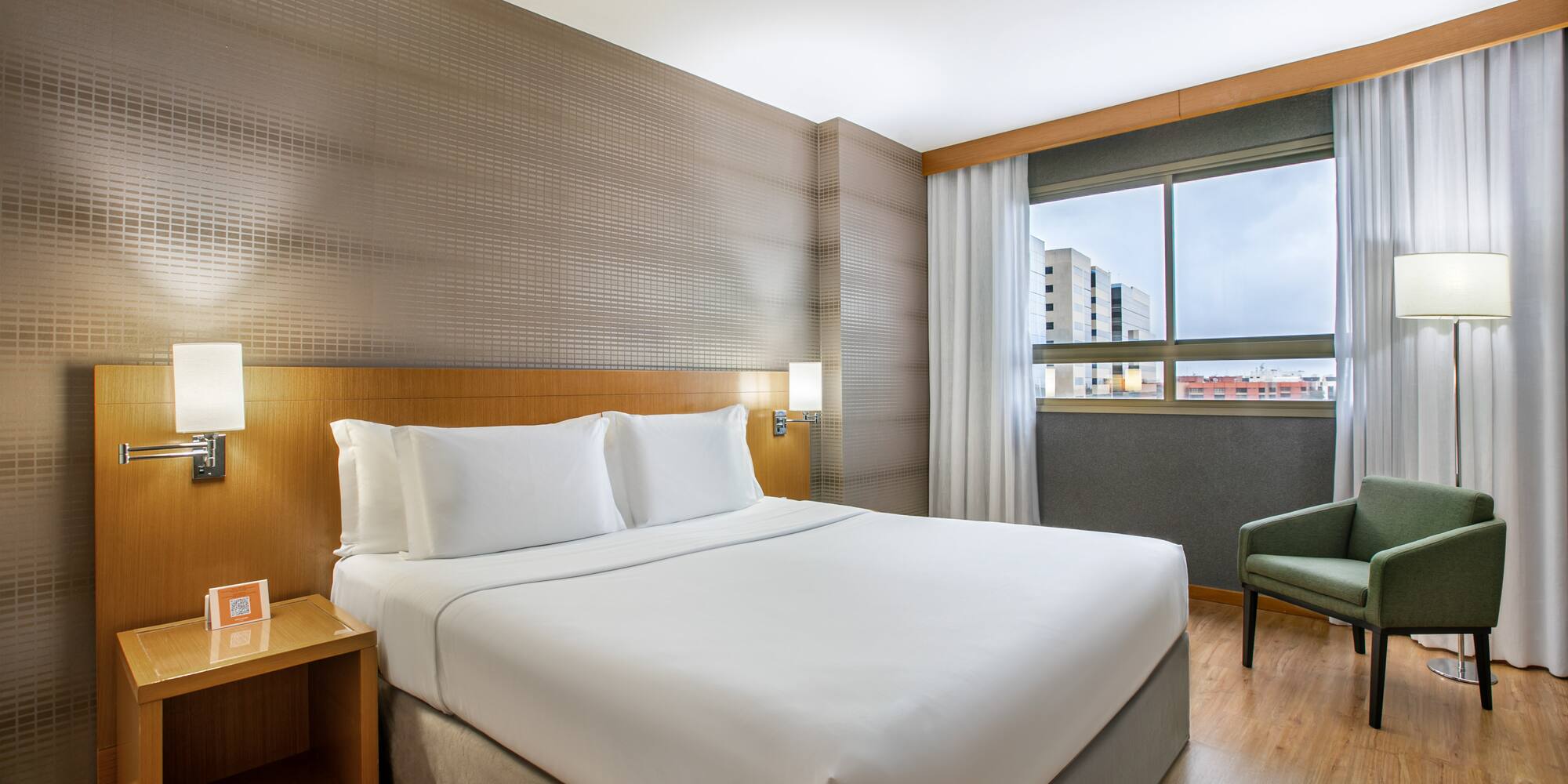 Modern hotel bedroom with king bed, green armchair, and window view.