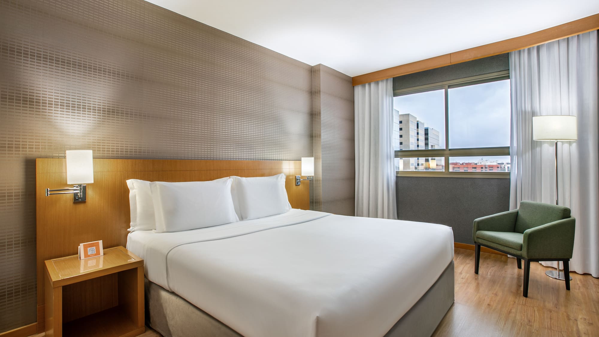 Modern hotel bedroom with king bed, green armchair, and window view.