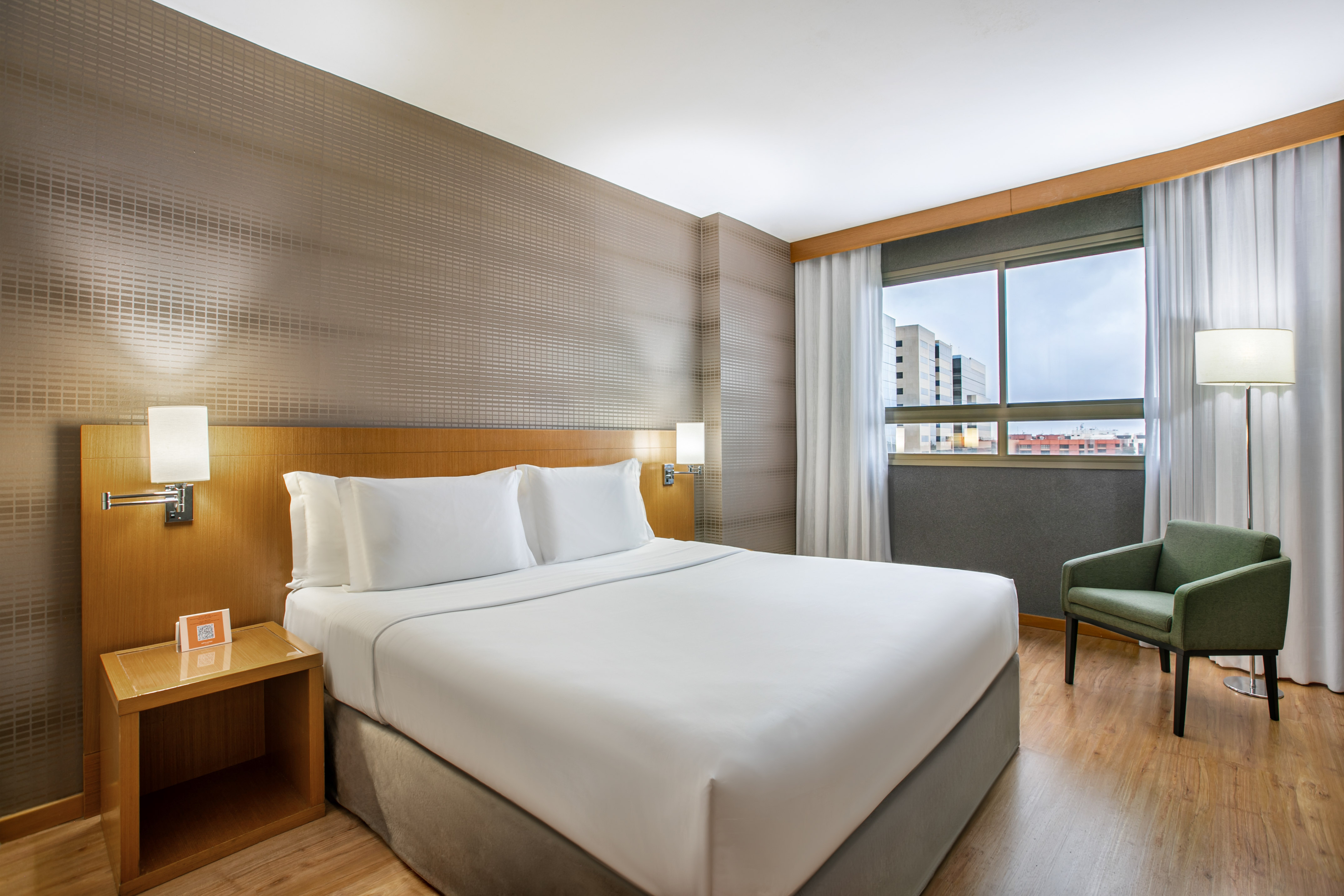 Modern hotel bedroom with king bed, green armchair, and window view.
