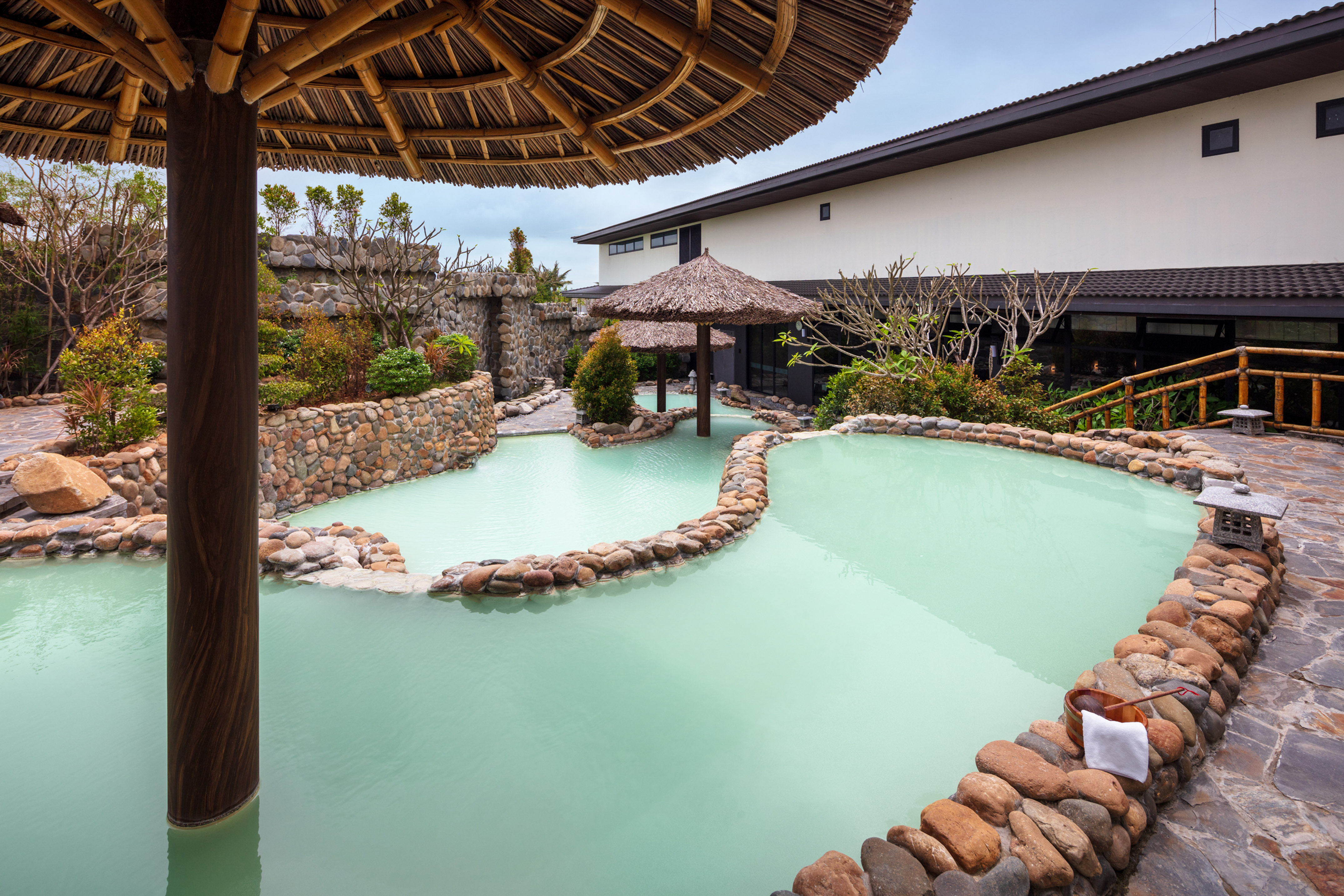 Outdoor onsen with light green water, rock walls, and thatched sunshades.