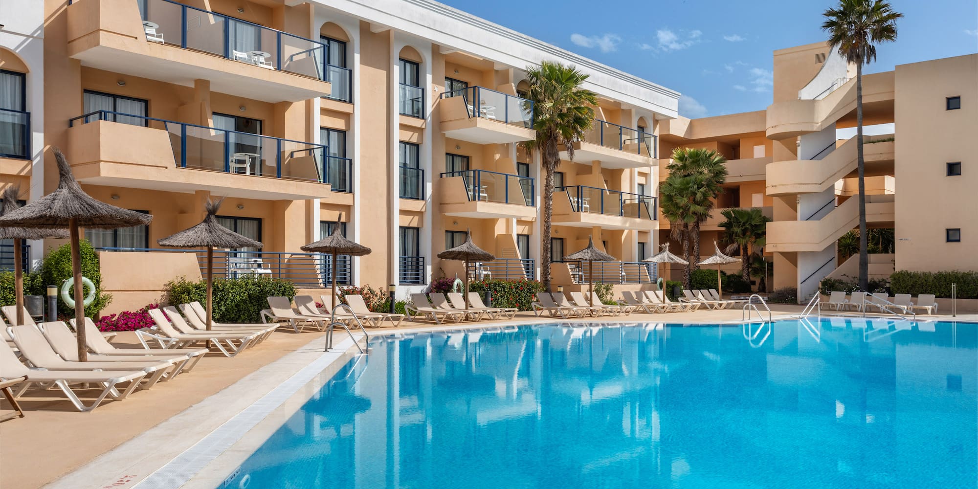 Sunny hotel resort with large outdoor swimming pool, palm trees, and lounge chairs.