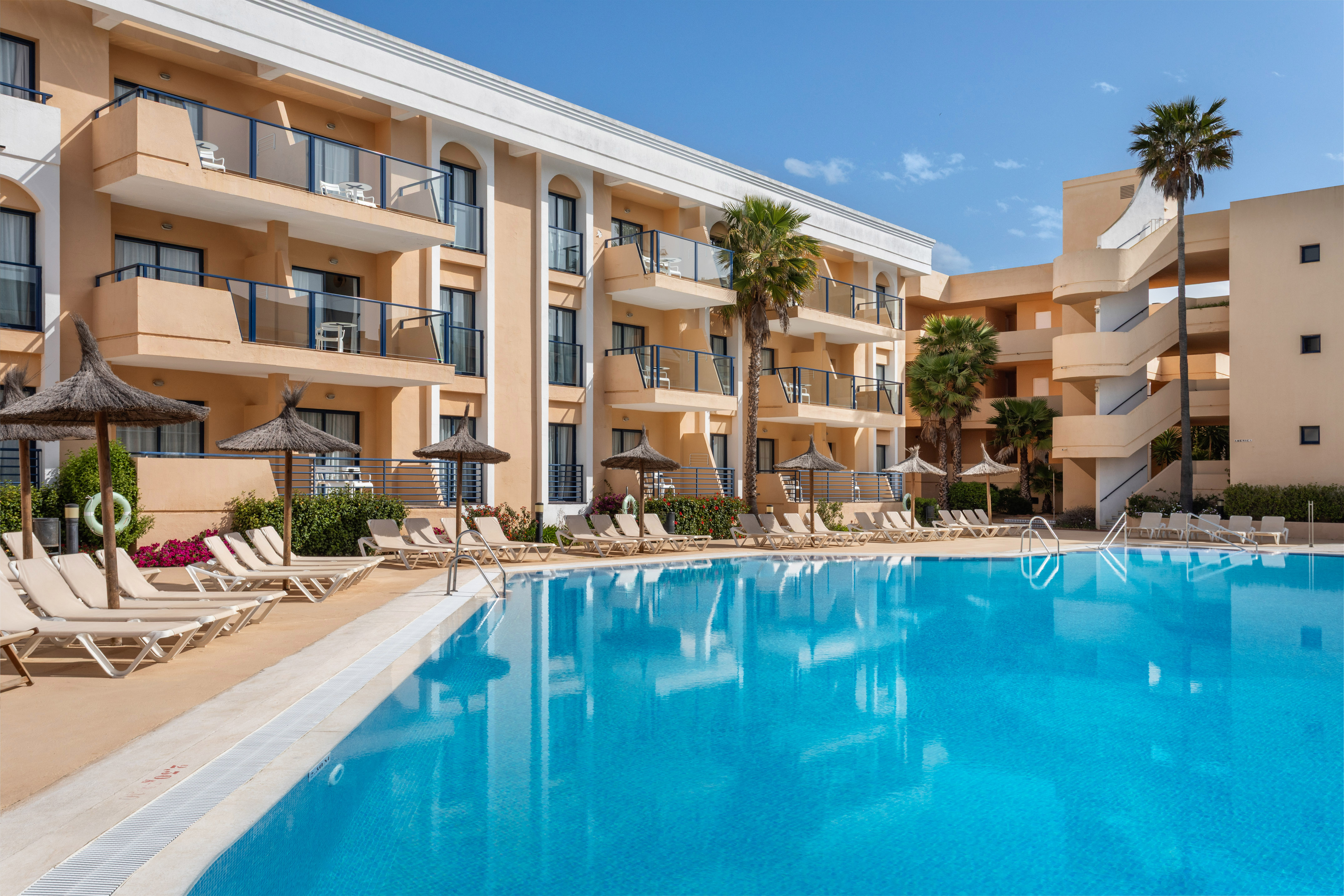 Sunny hotel resort with large outdoor swimming pool, palm trees, and lounge chairs.