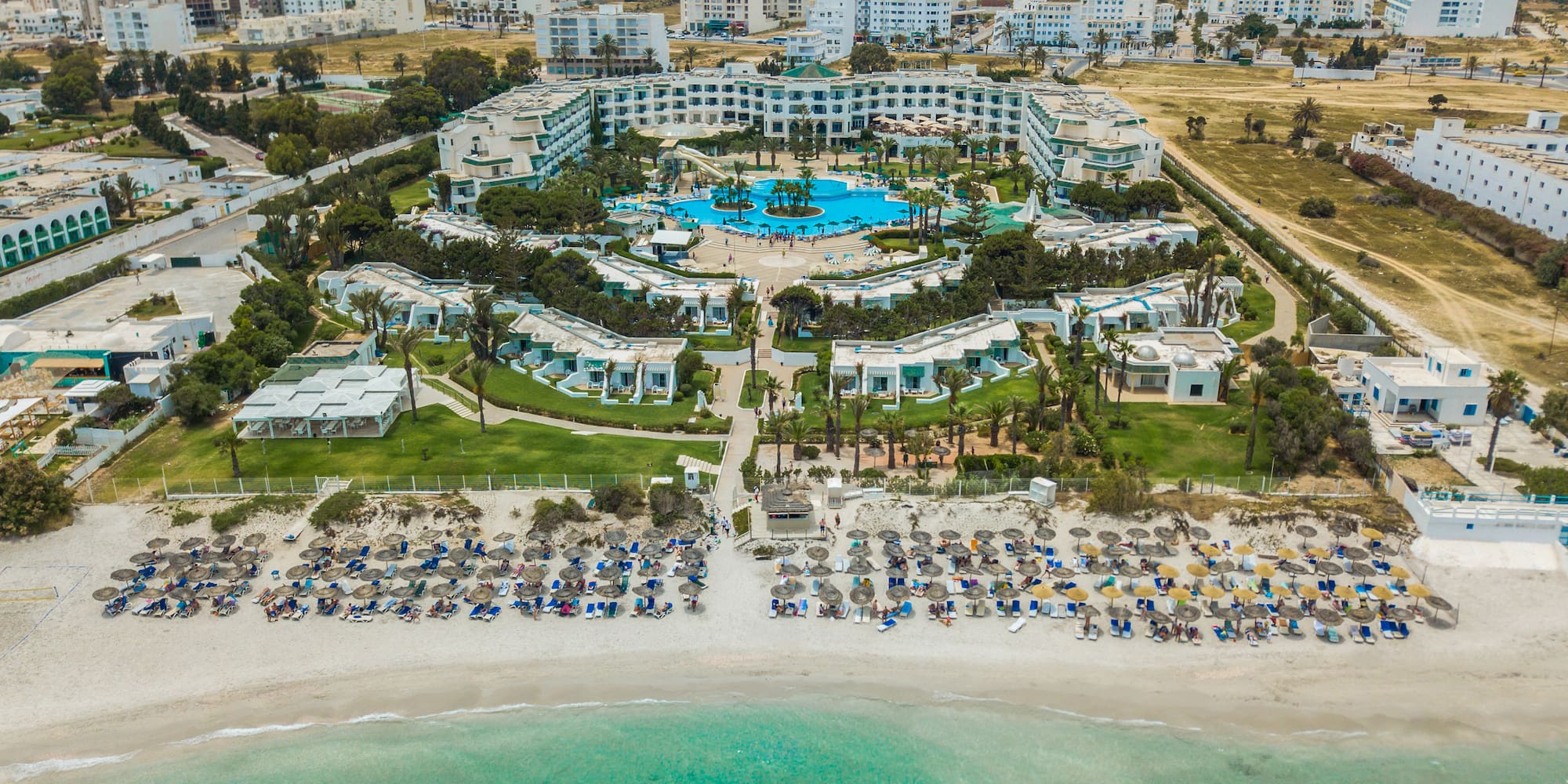 Aerial beach resort hotel with large pool, palm trees, and turquoise sea.