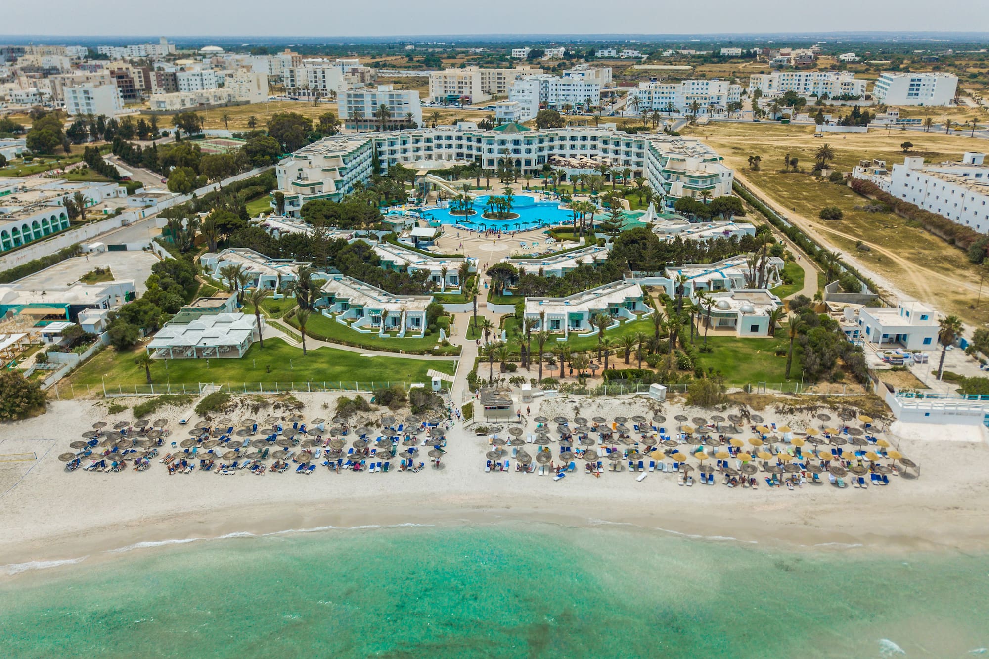 Aerial beach resort hotel with large pool, palm trees, and turquoise sea.