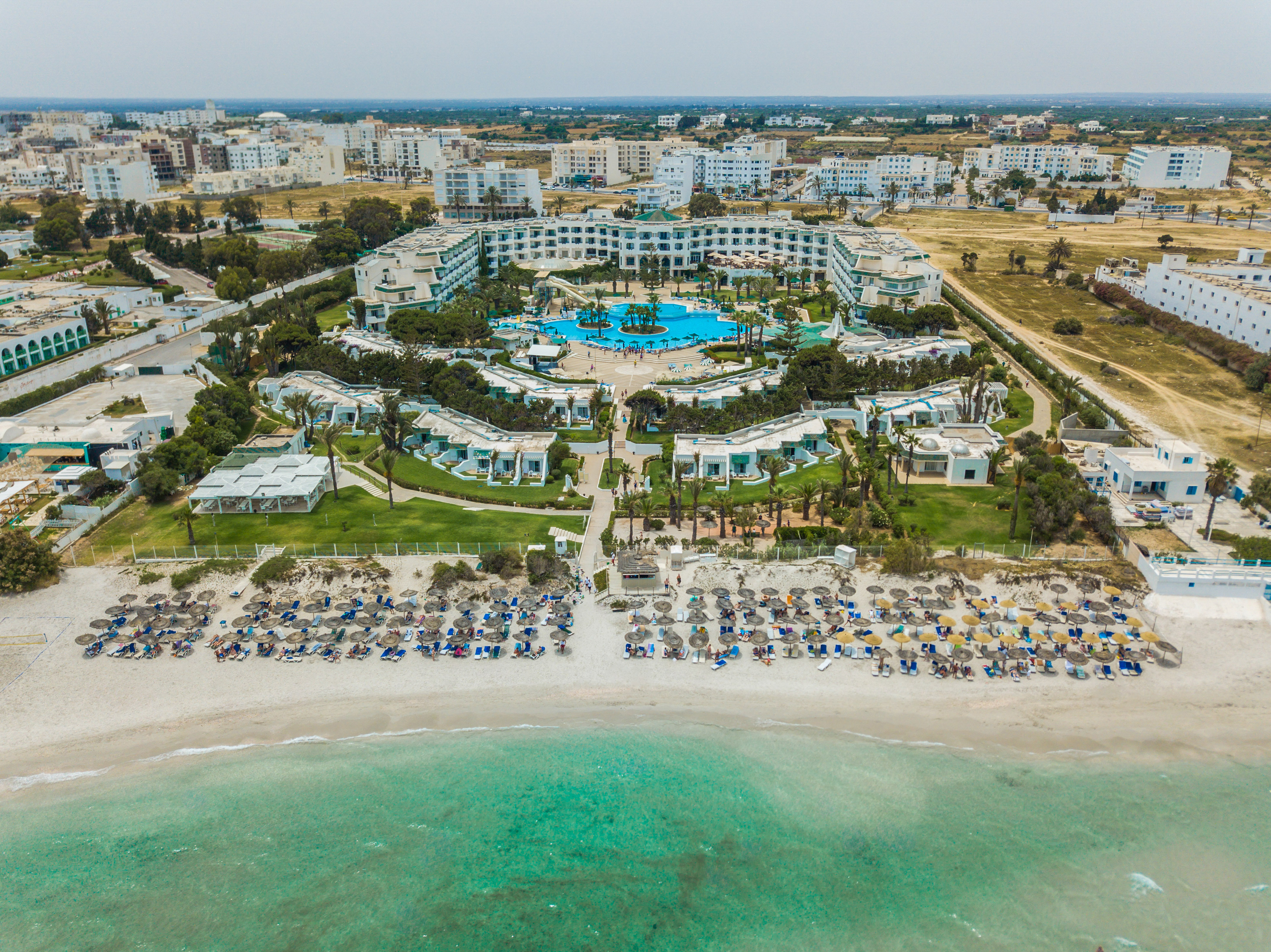 Aerial beach resort hotel with large pool, palm trees, and turquoise sea.