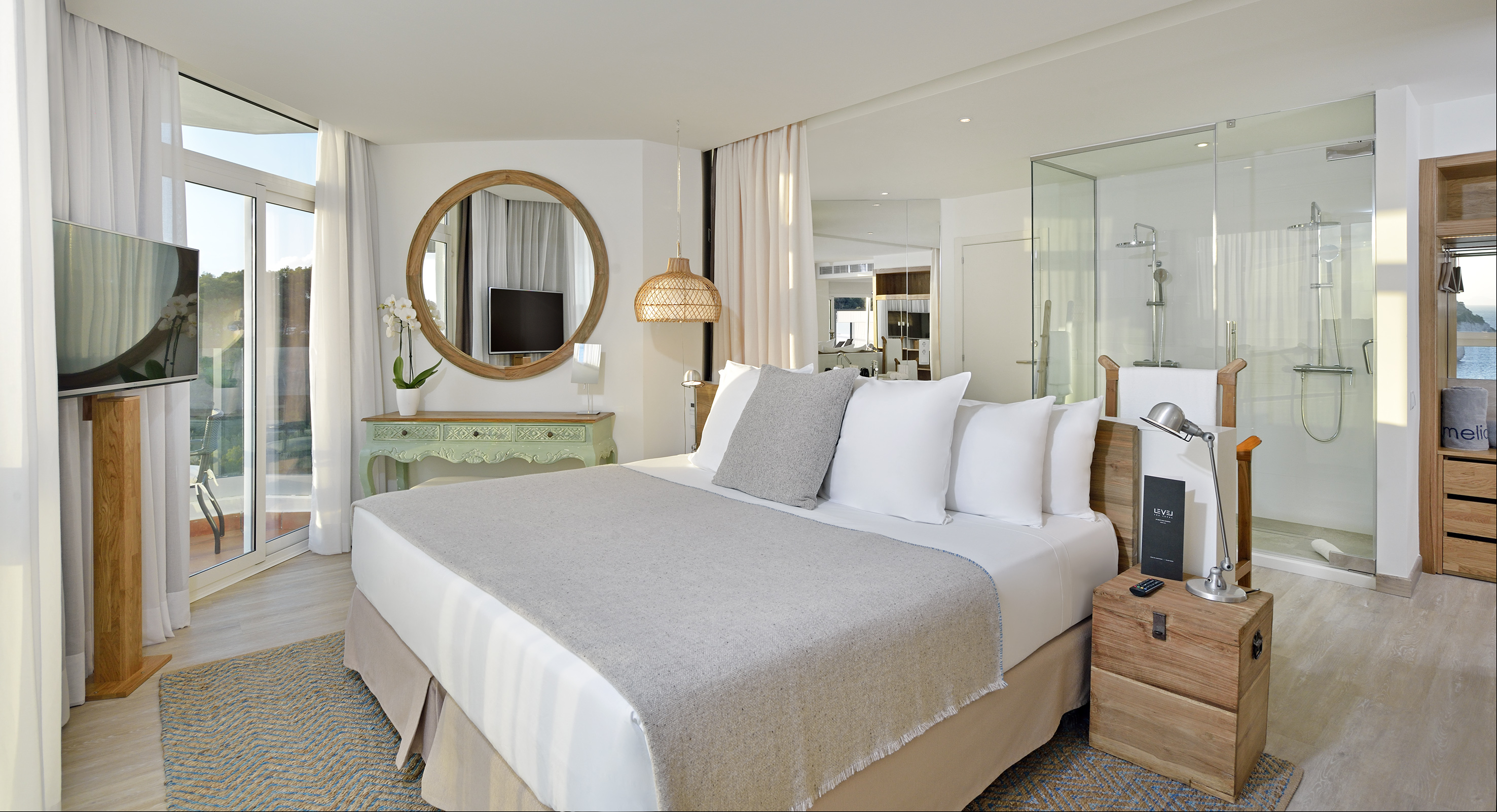 Bright modern hotel bedroom with sea view balcony and open glass shower.