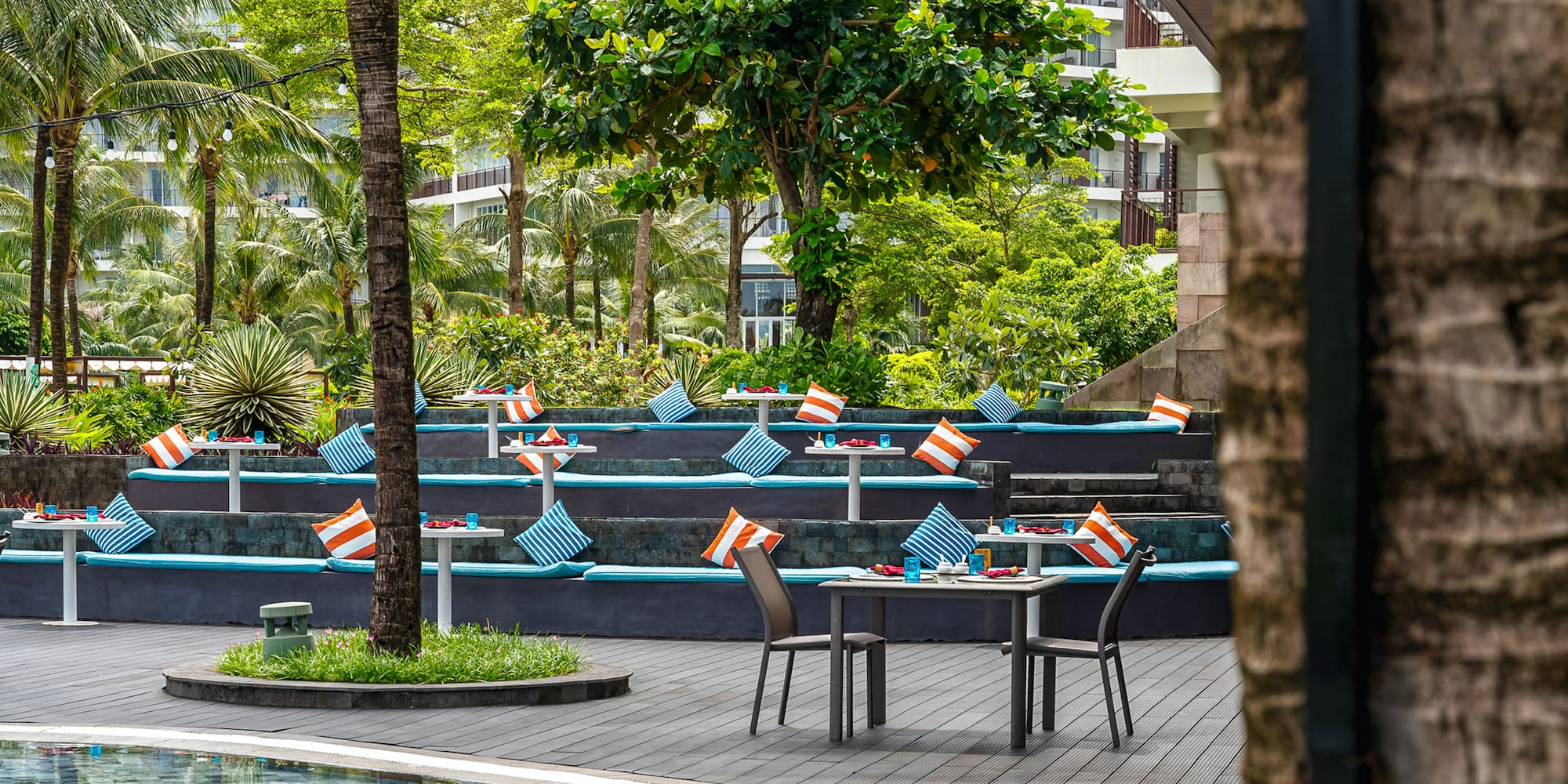 Poolside tropical resort dining with tiered seating, blue cushions, and palm trees.