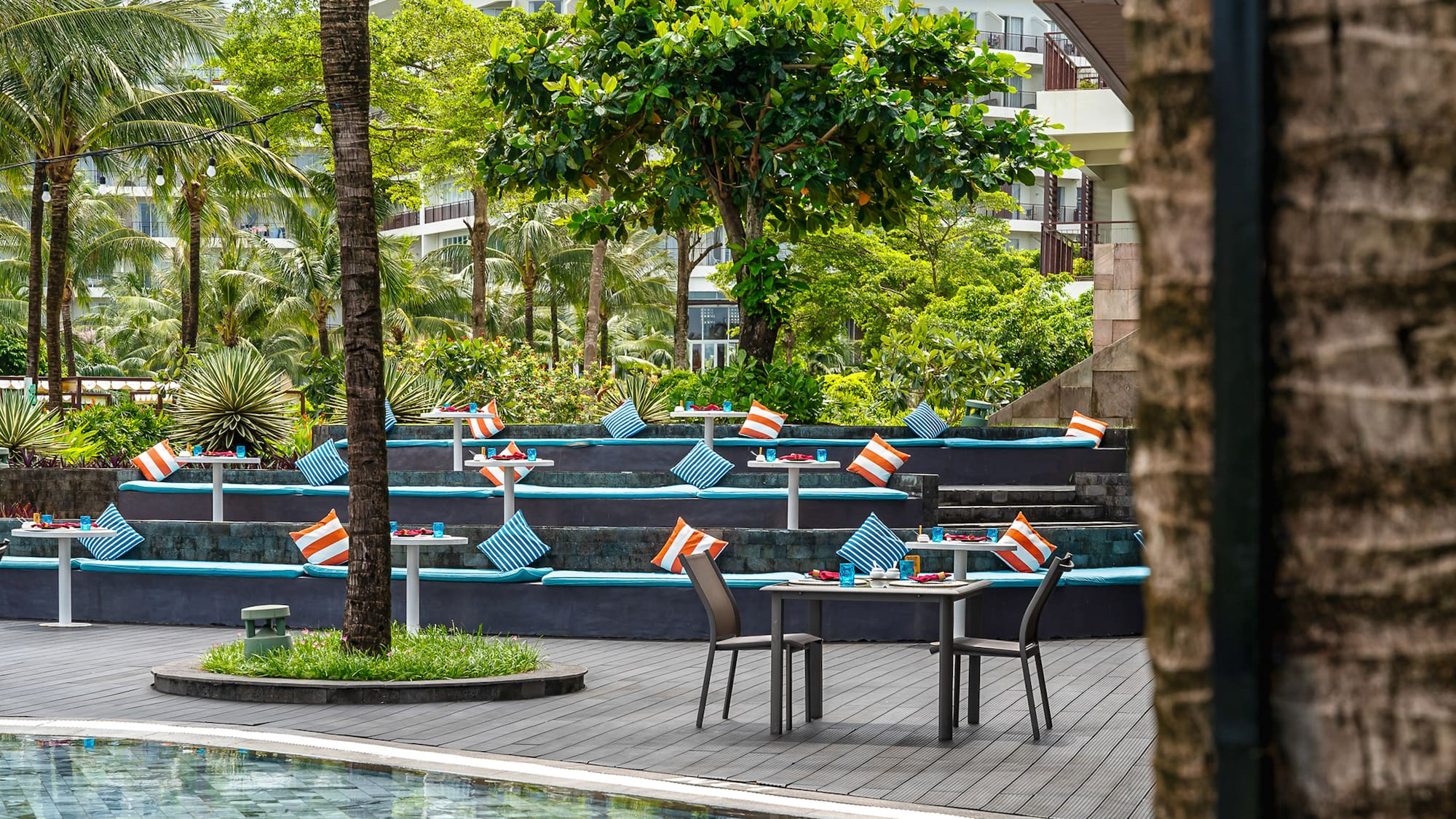 Poolside tropical resort dining with tiered seating, blue cushions, and palm trees.
