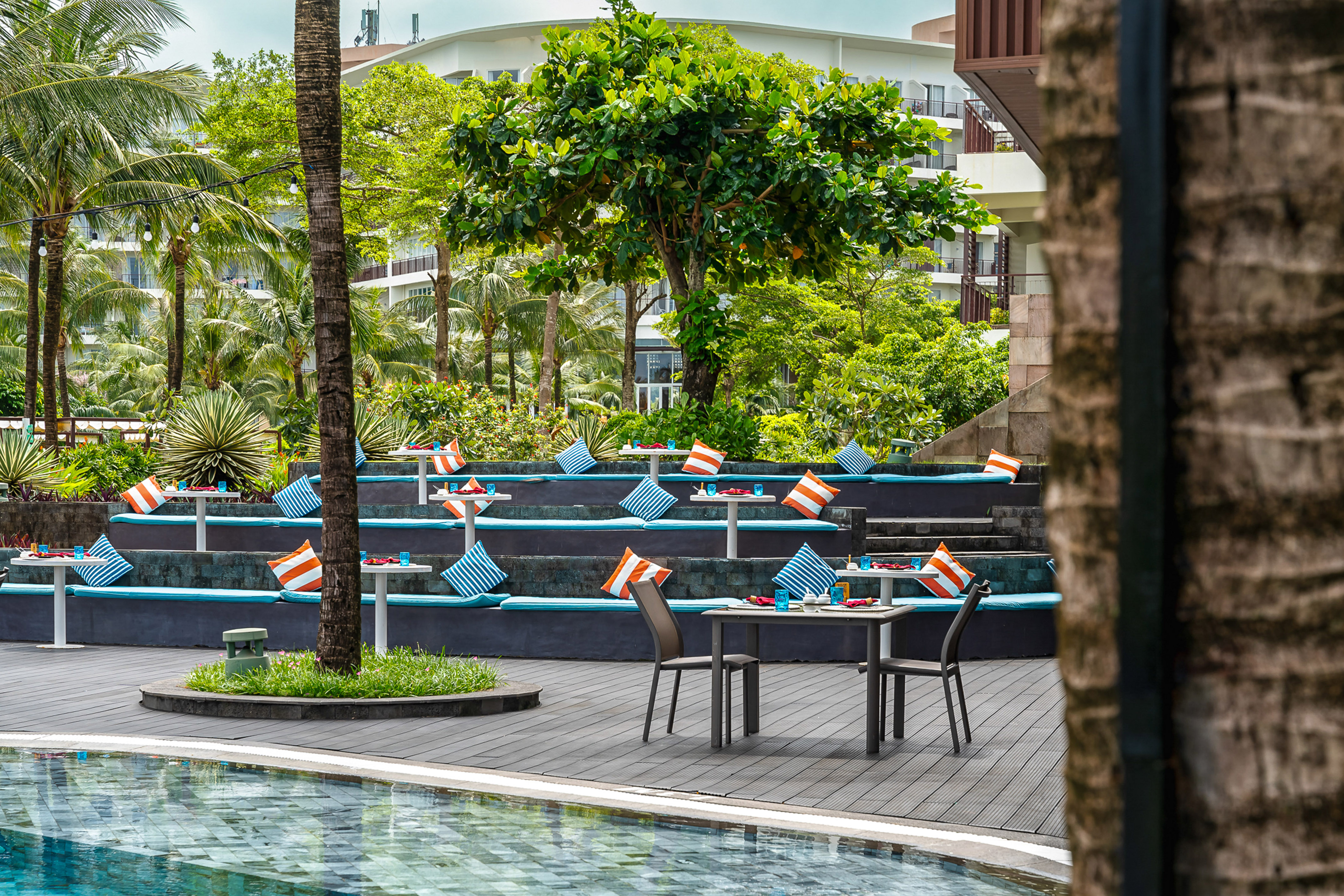 Poolside tropical resort dining with tiered seating, blue cushions, and palm trees.