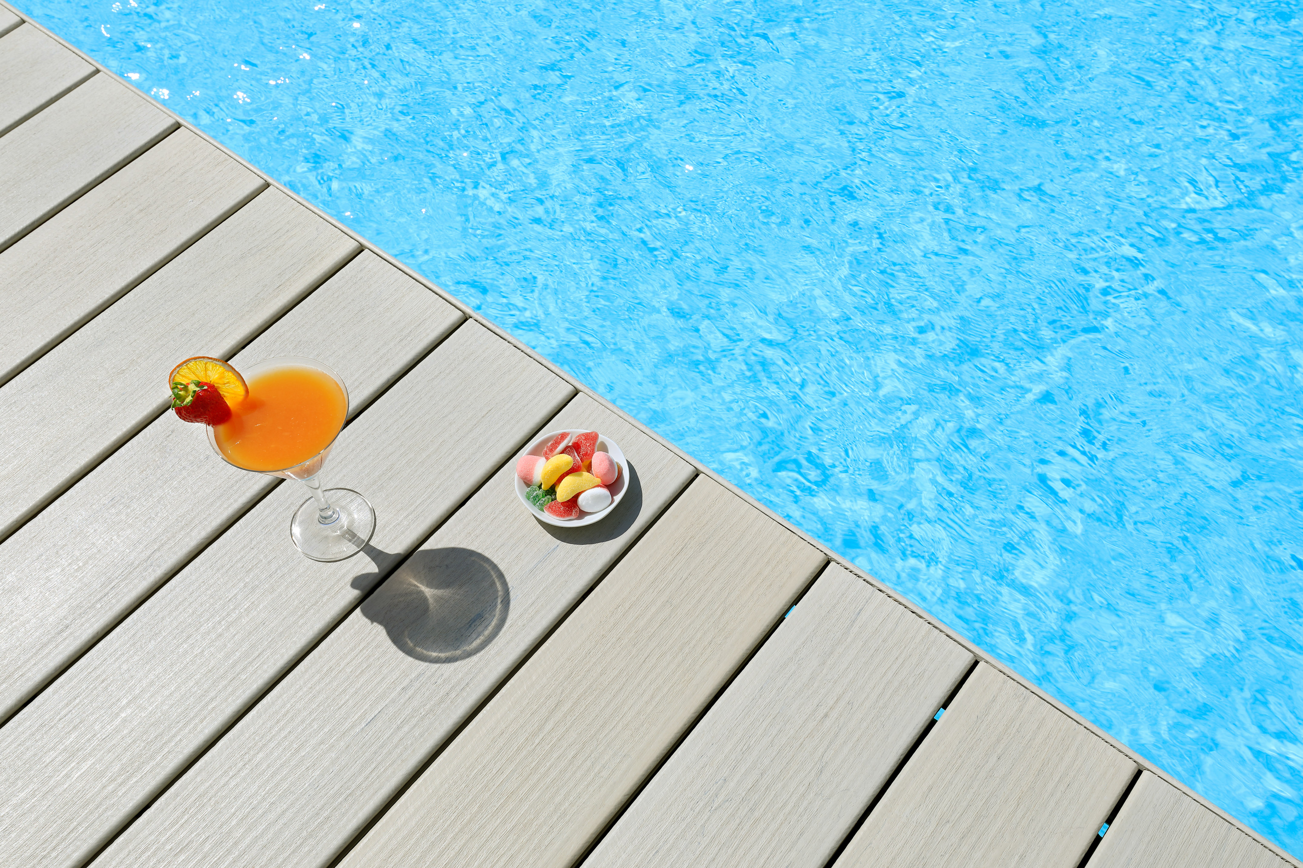 Orange cocktail, colorful candies on poolside deck by blue swimming pool.