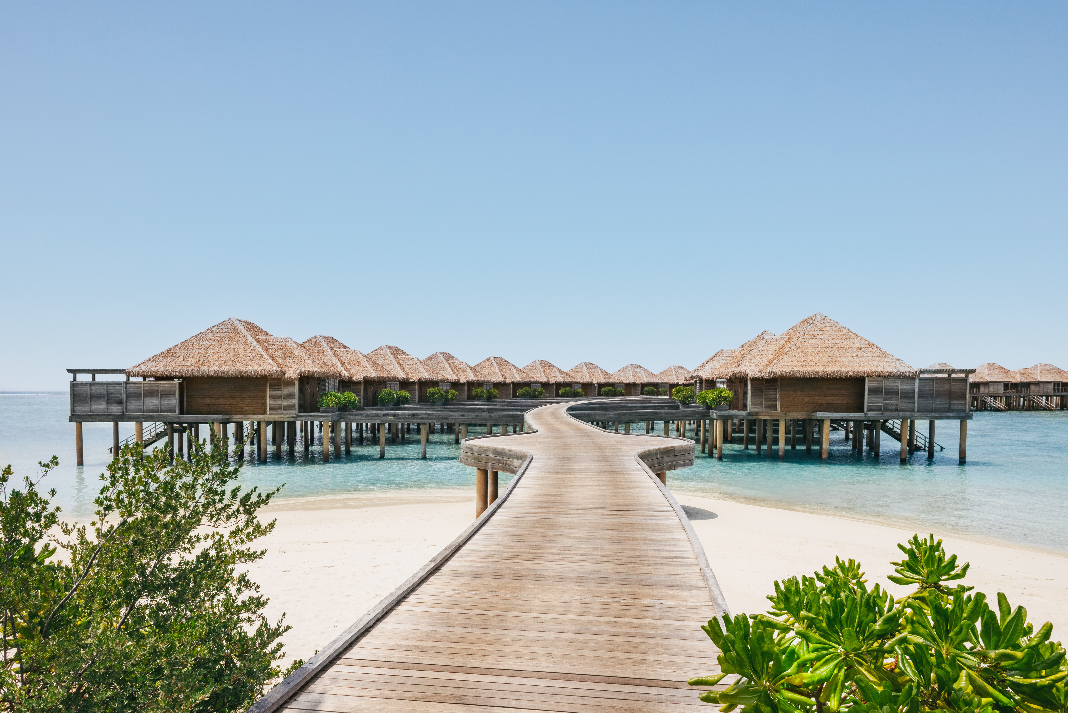 Luxury overwater bungalows, thatched roofs, wooden boardwalk, clear blue water.