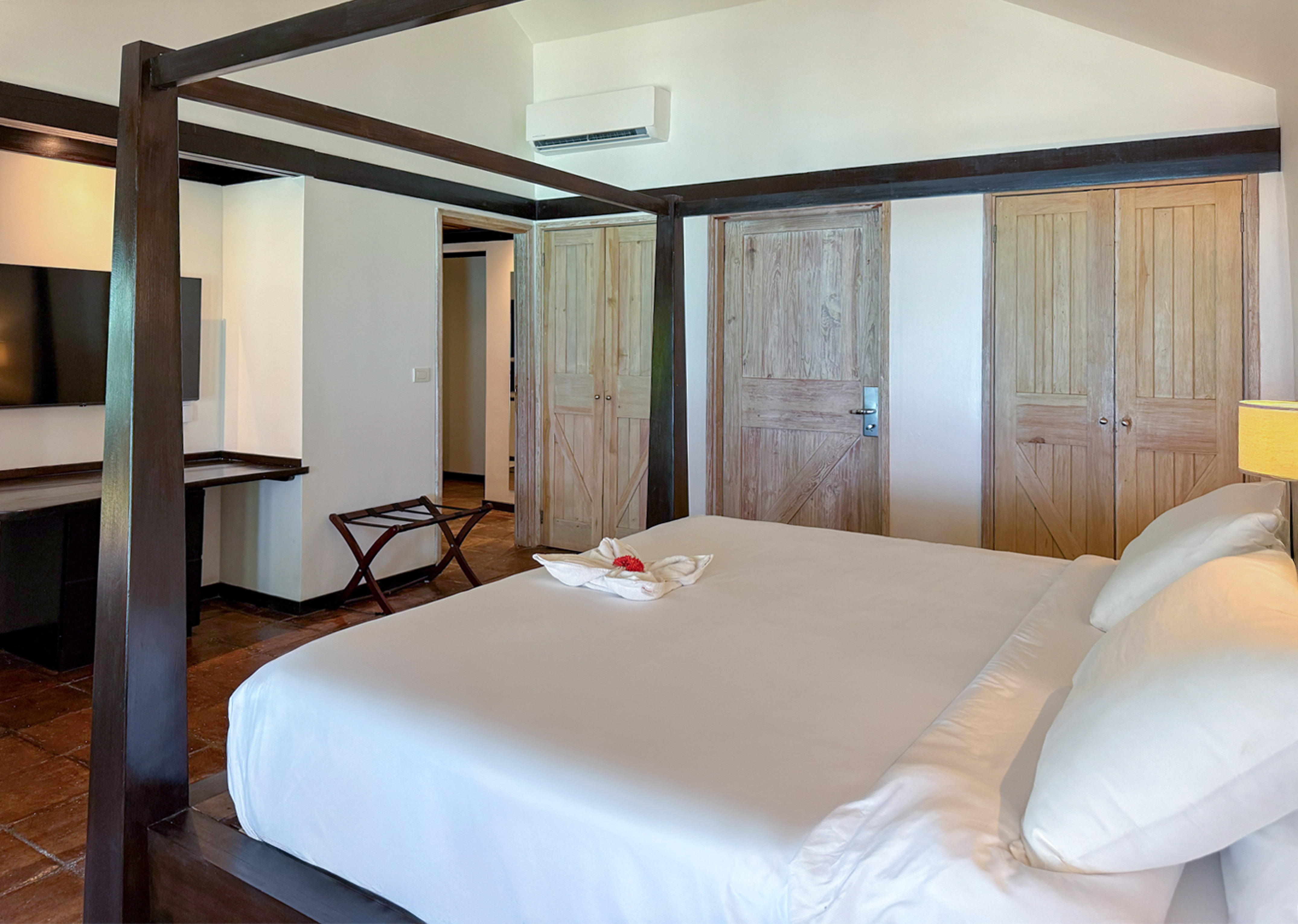 Tropical hotel bedroom with dark four-poster bed, white bedding, wooden doors.