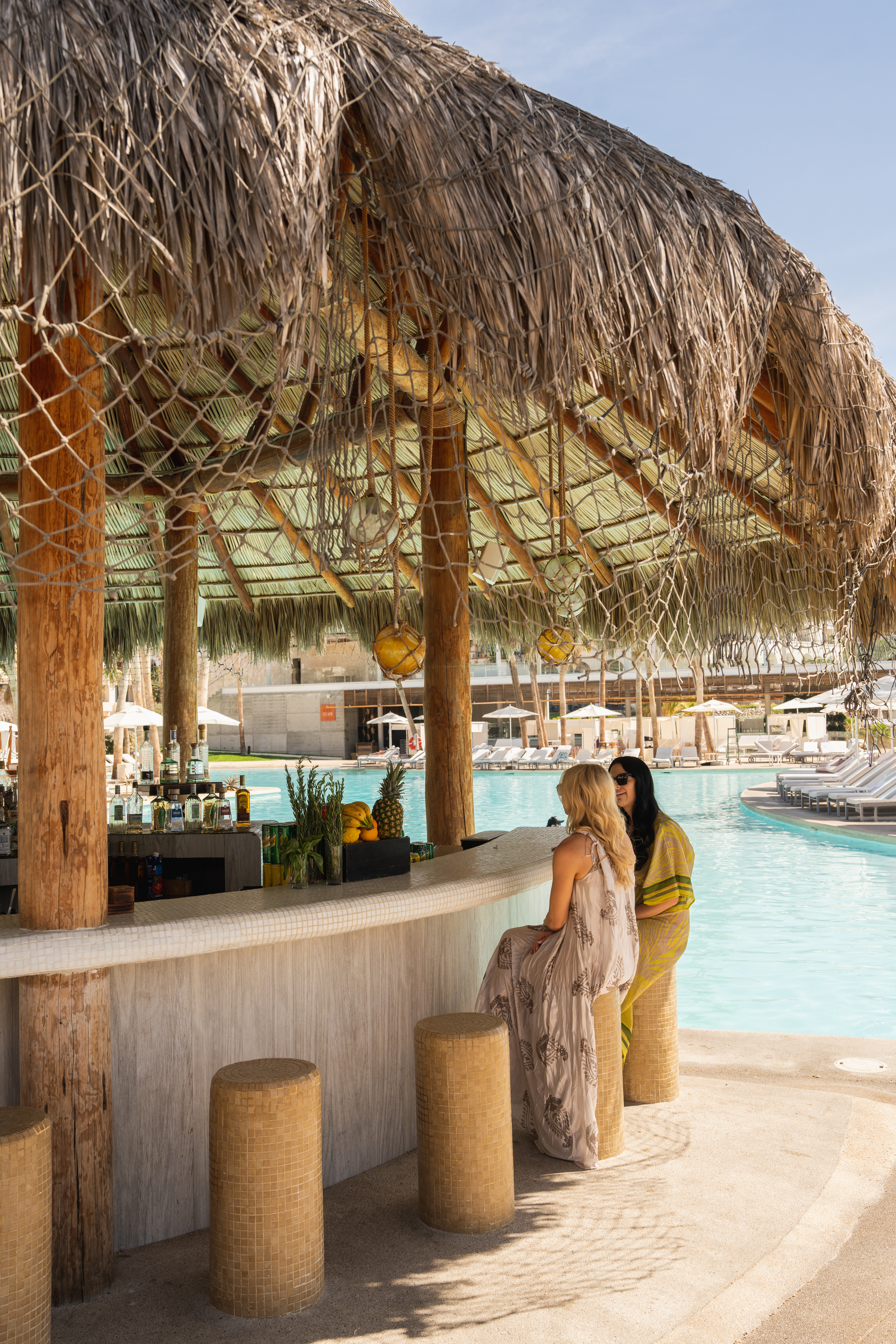 Two women at a tropical resort poolside bar with a thatched palapa roof.