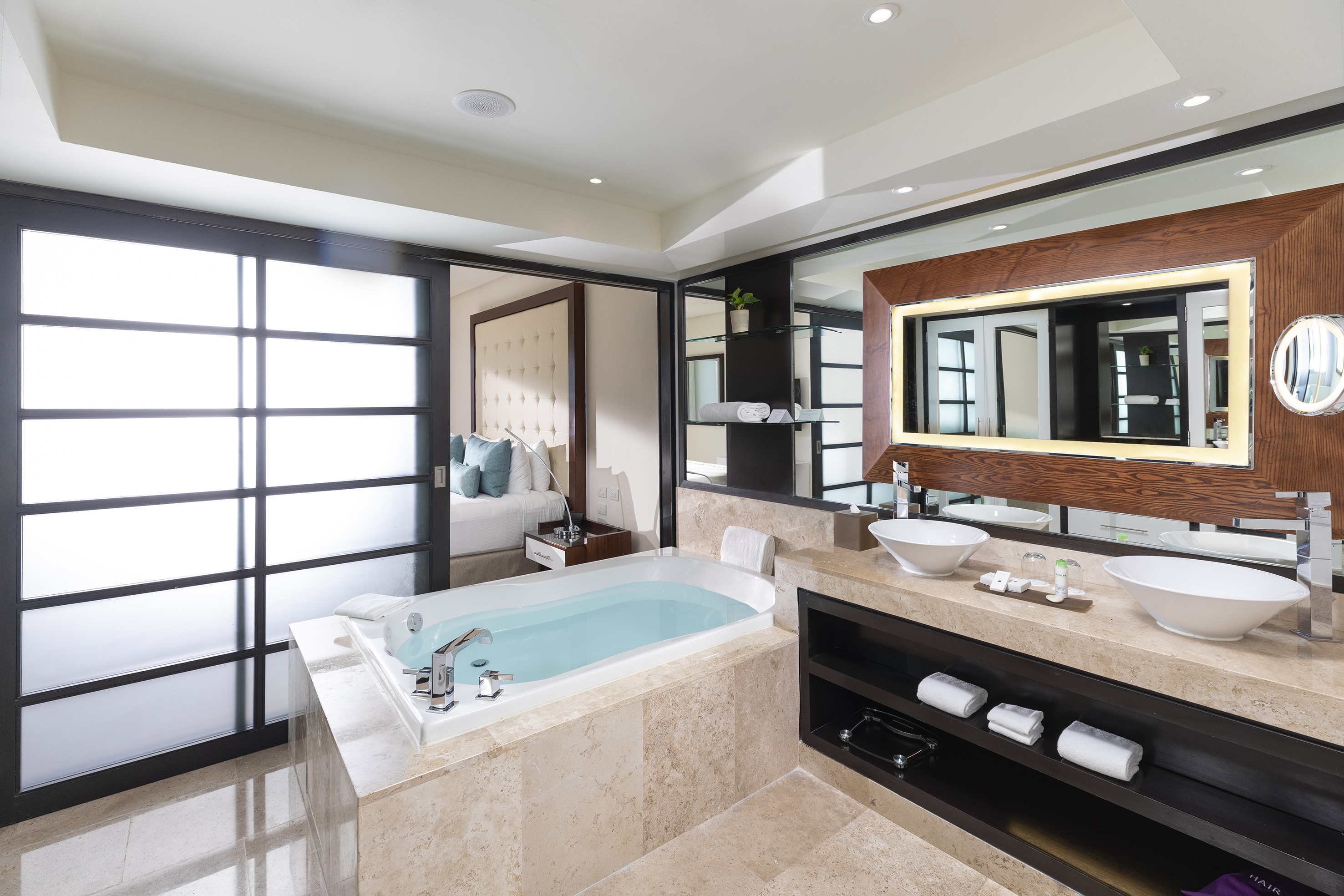 Modern hotel bathroom with soaking tub, dual vanity, and adjacent bedroom.