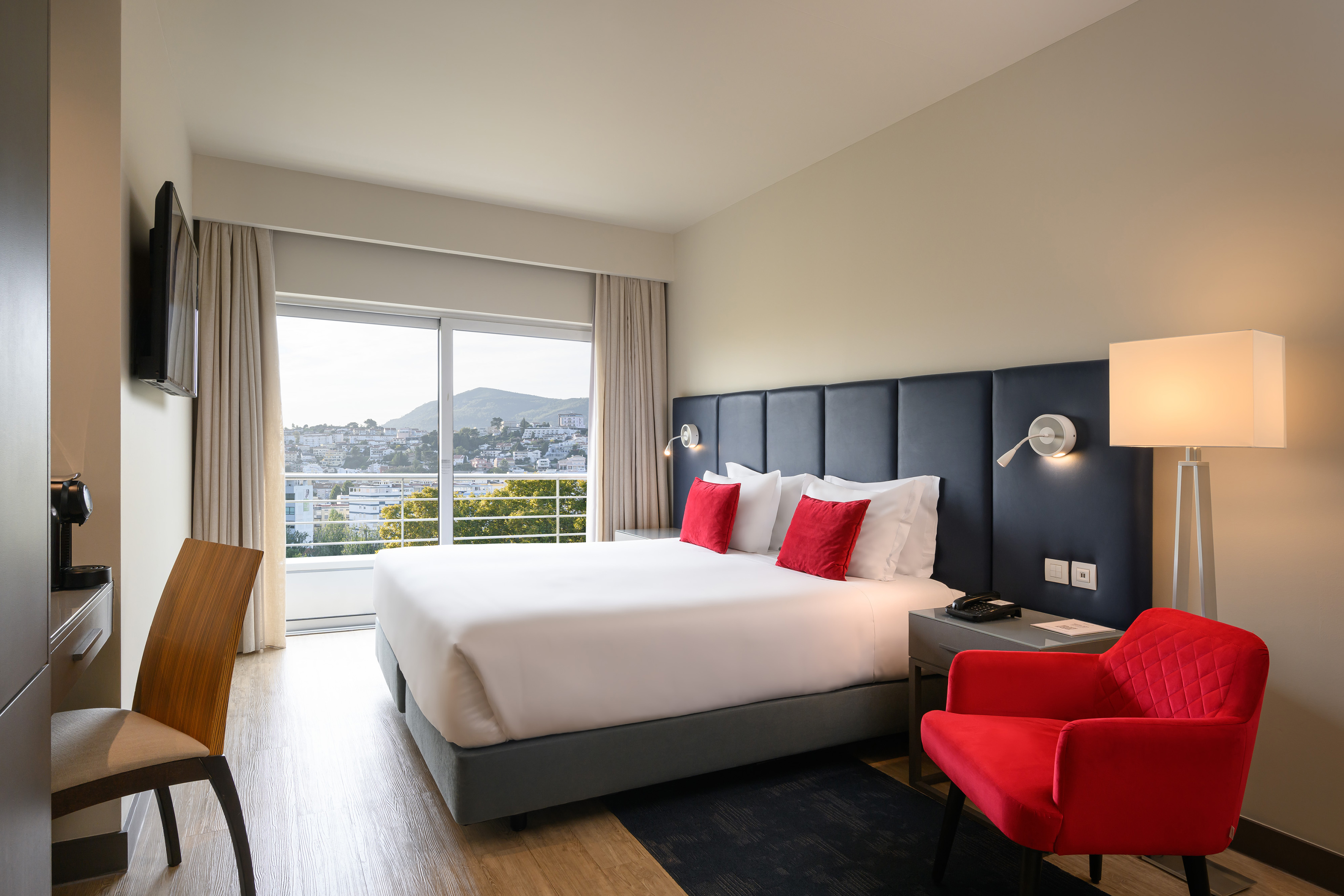 Modern hotel room with king bed, red armchair, and large window city view.