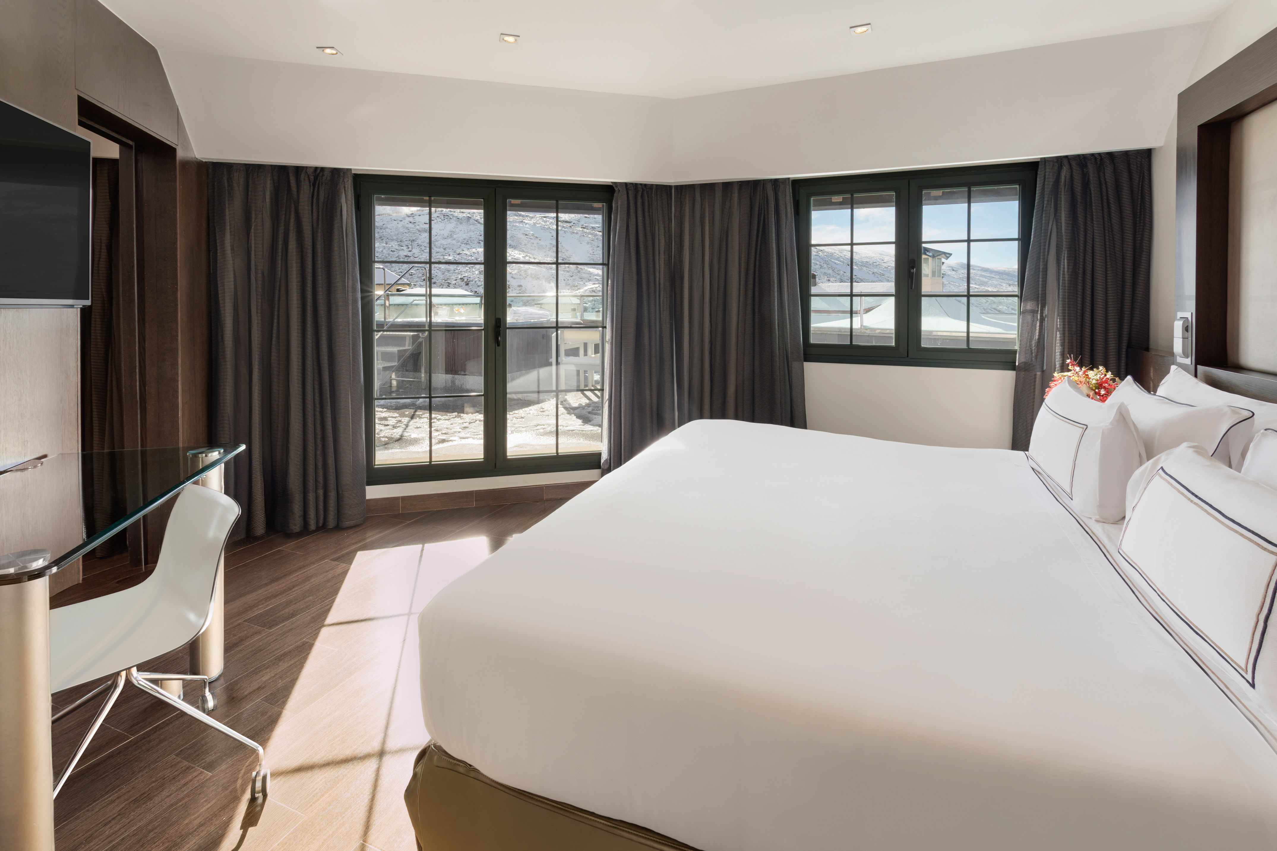 Modern hotel room with bed, desk, and scenic snowy mountain view.