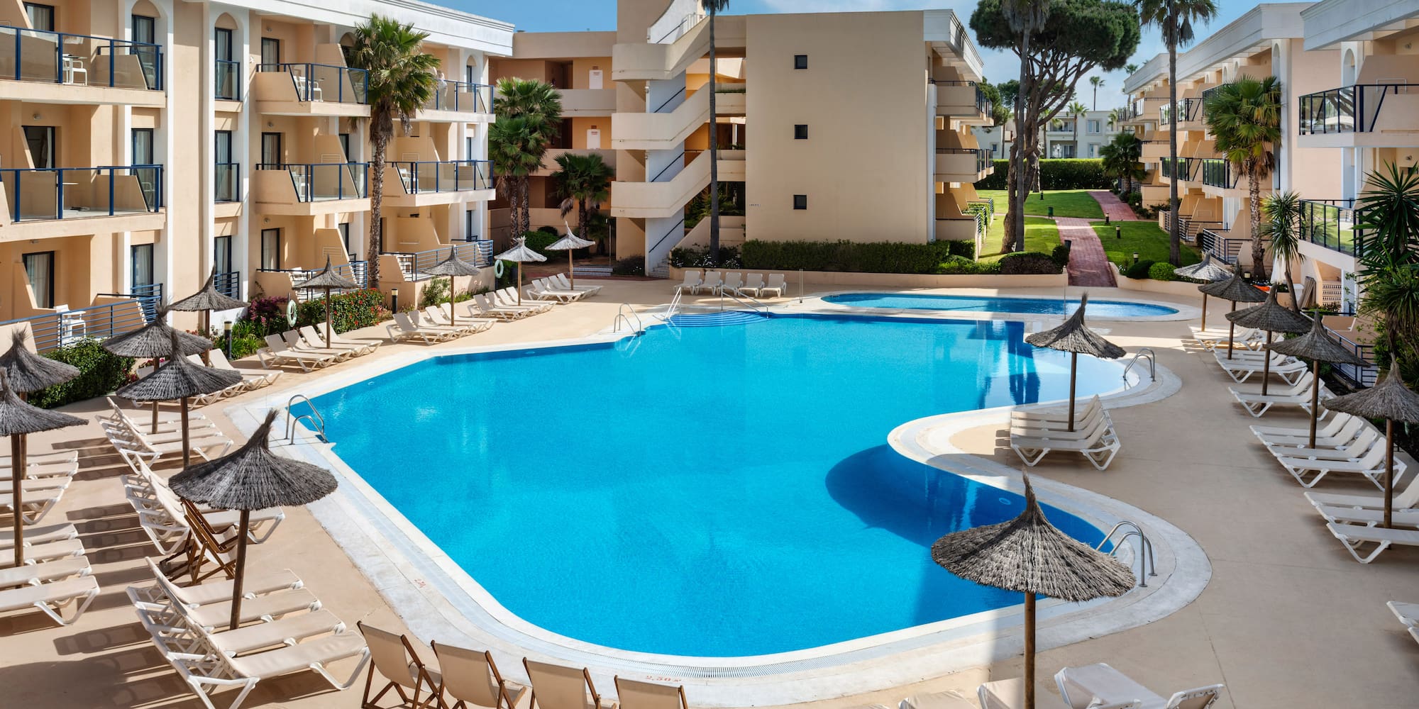Outdoor hotel pool, sun loungers, palm trees, and resort buildings.