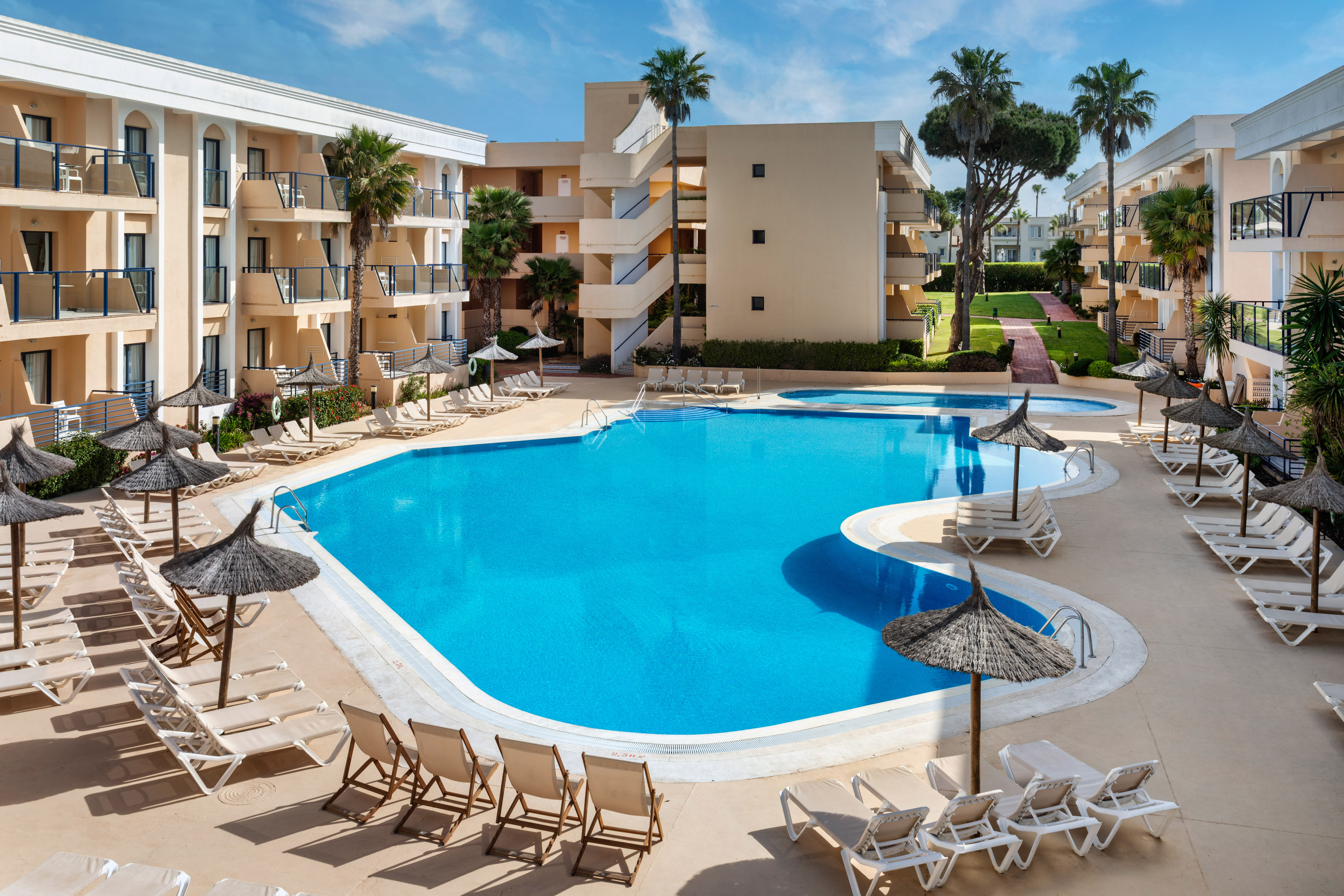 Gran piscina de hotel con tumbonas y parasoles, rodeada de edificios y palmeras.