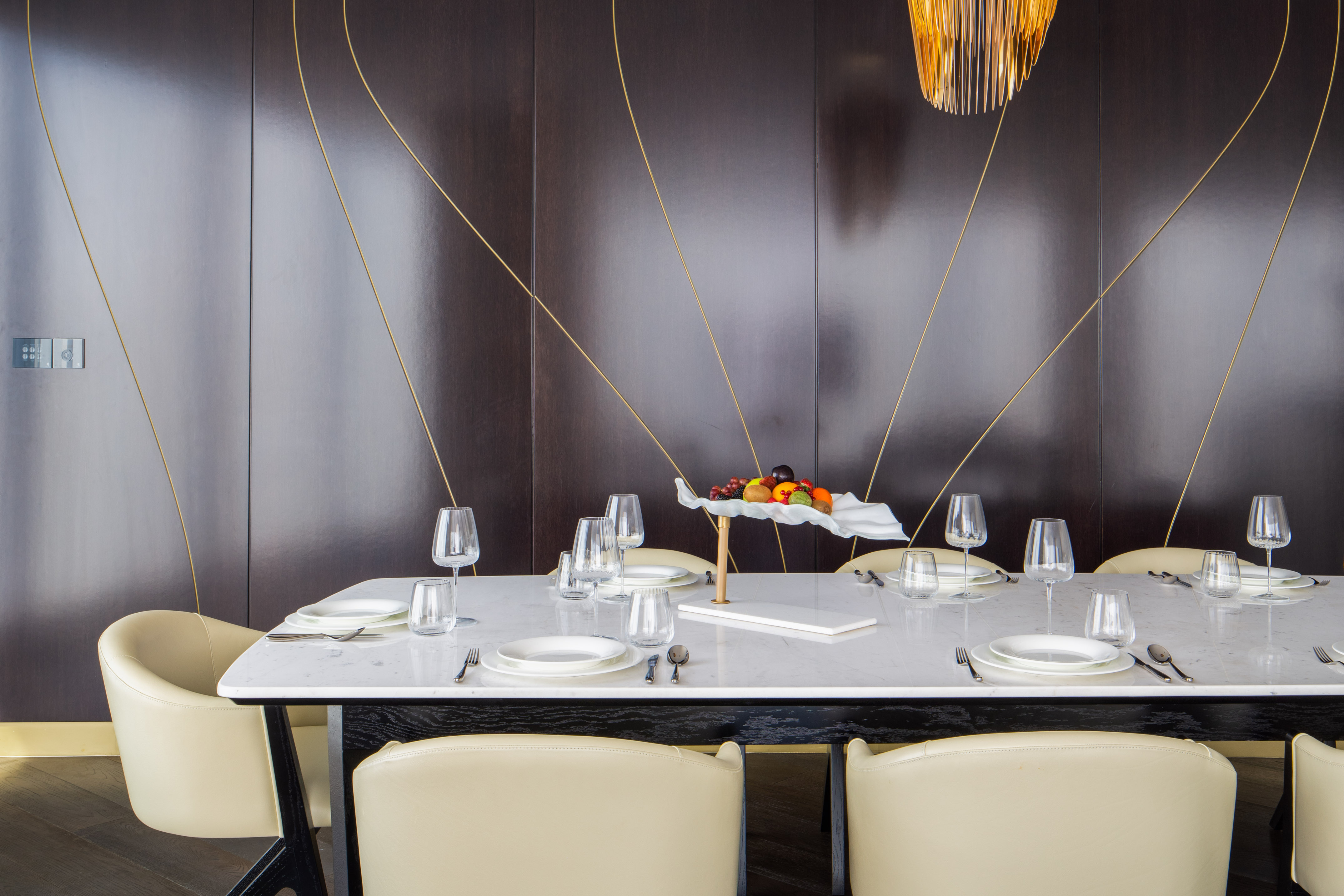 Elegant dining room with marble table, cream chairs, dark wood and gold wall, fruit bowl.