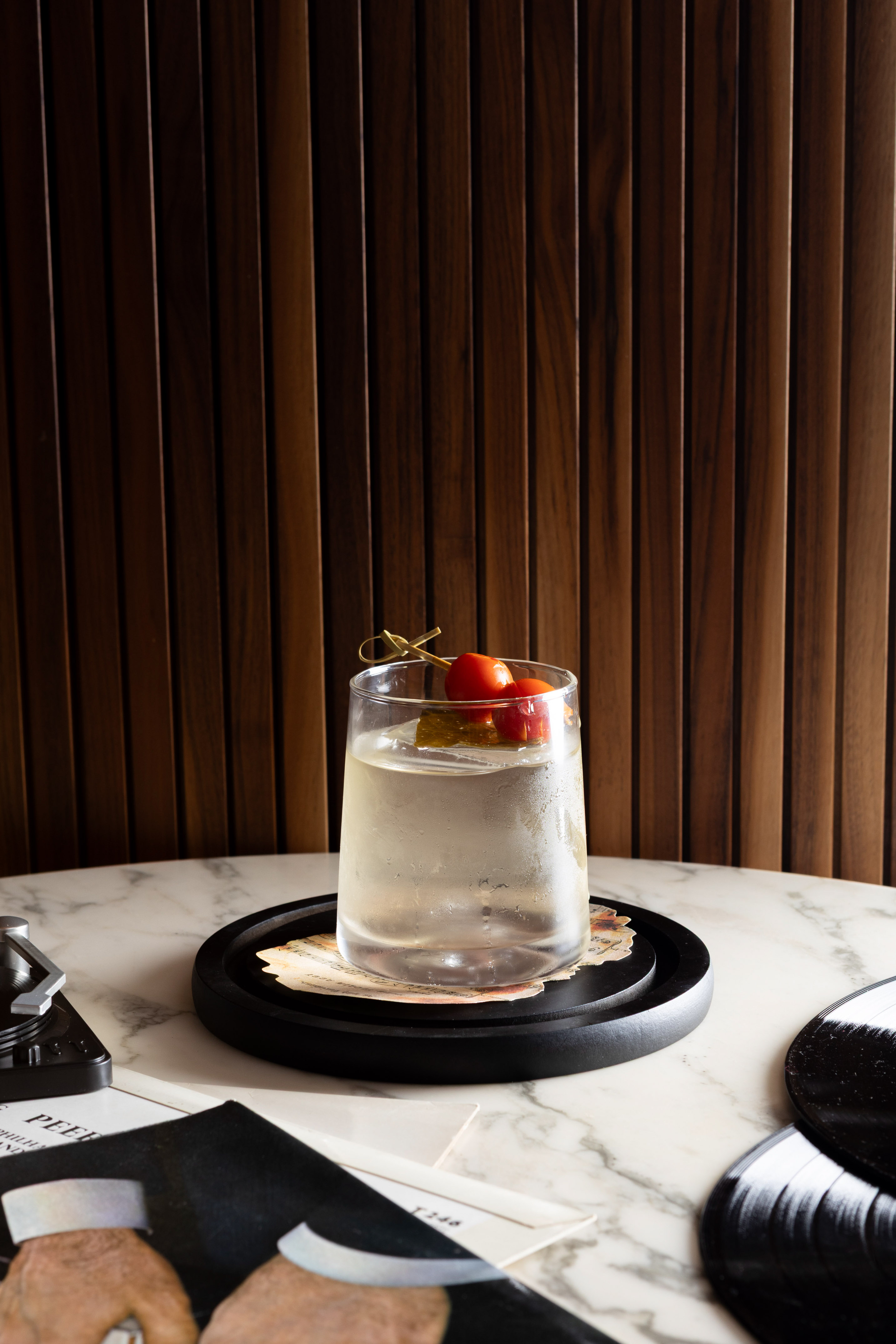 Clear cocktail with cherry tomato, pickle garnish, marble table, vinyl records.
