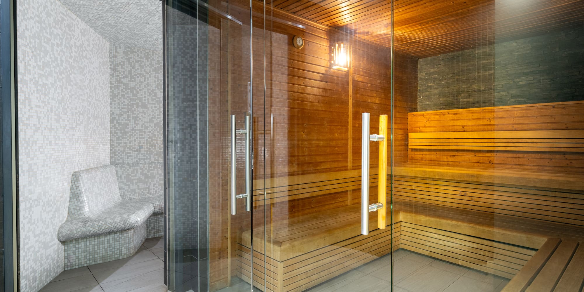 Modern wooden sauna and adjacent mosaic steam room with glass door.