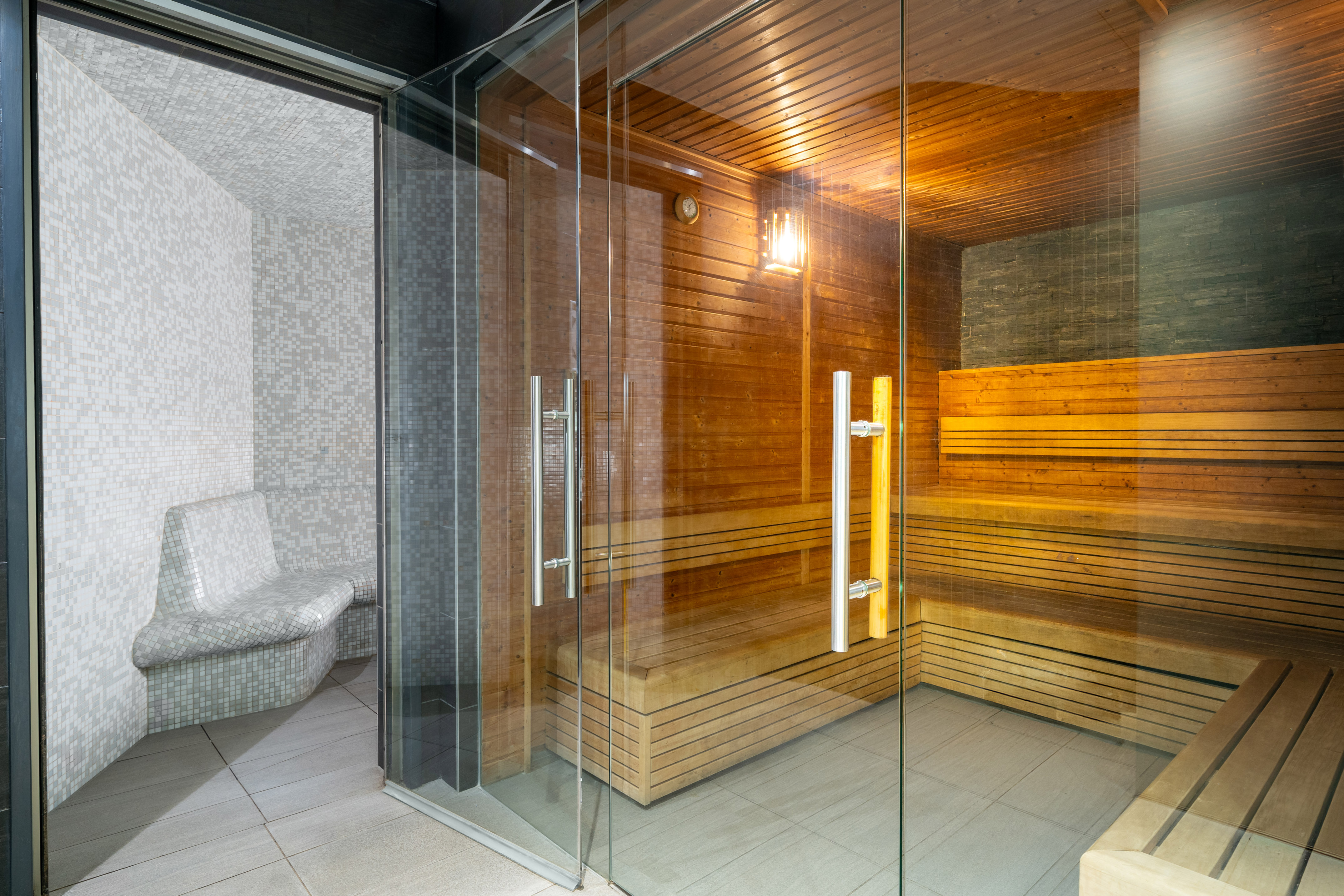 Modern wooden sauna and adjacent mosaic steam room with glass door.