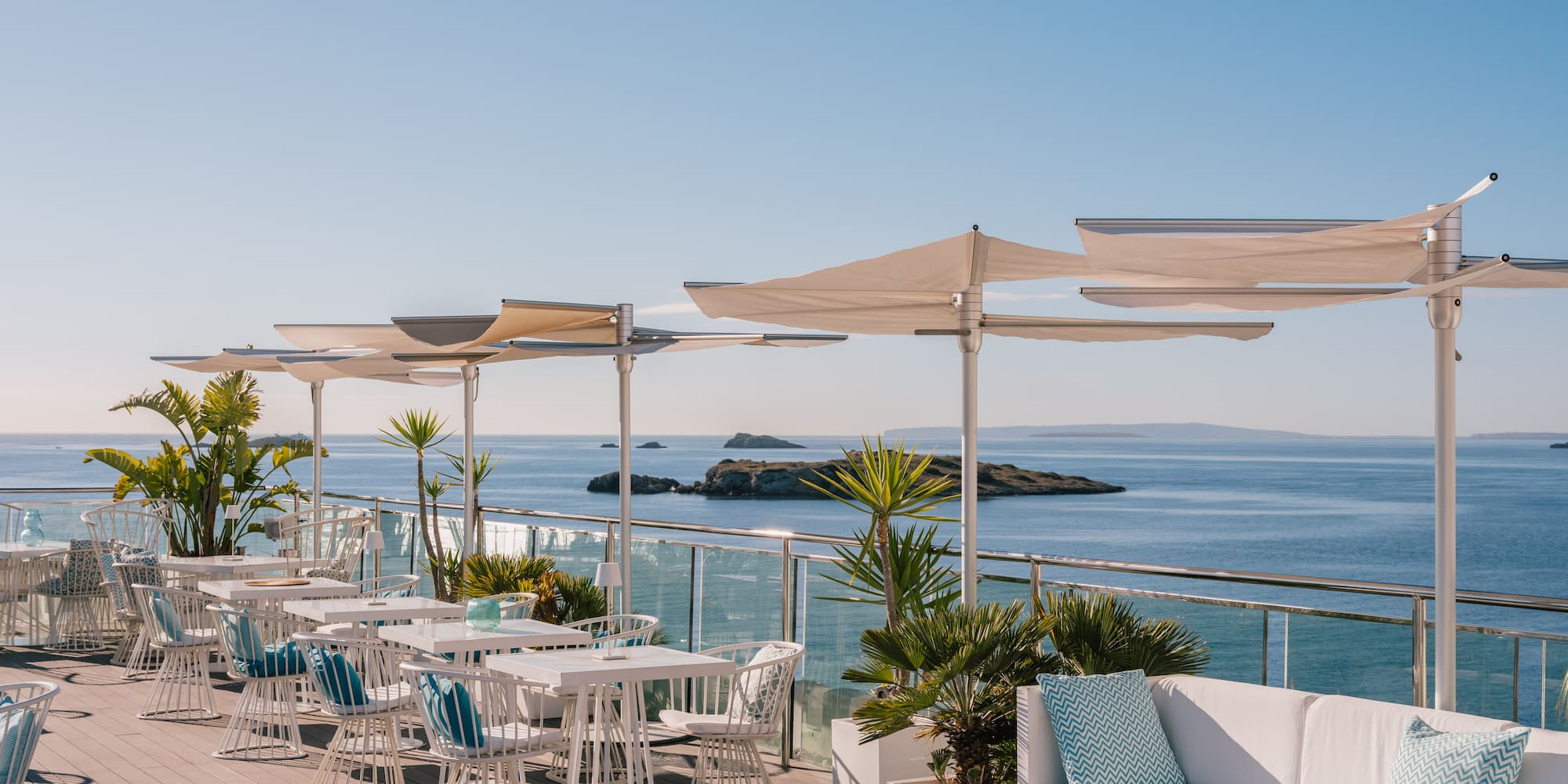 Sunny luxury rooftop terrace overlooking ocean, white outdoor dining furniture.