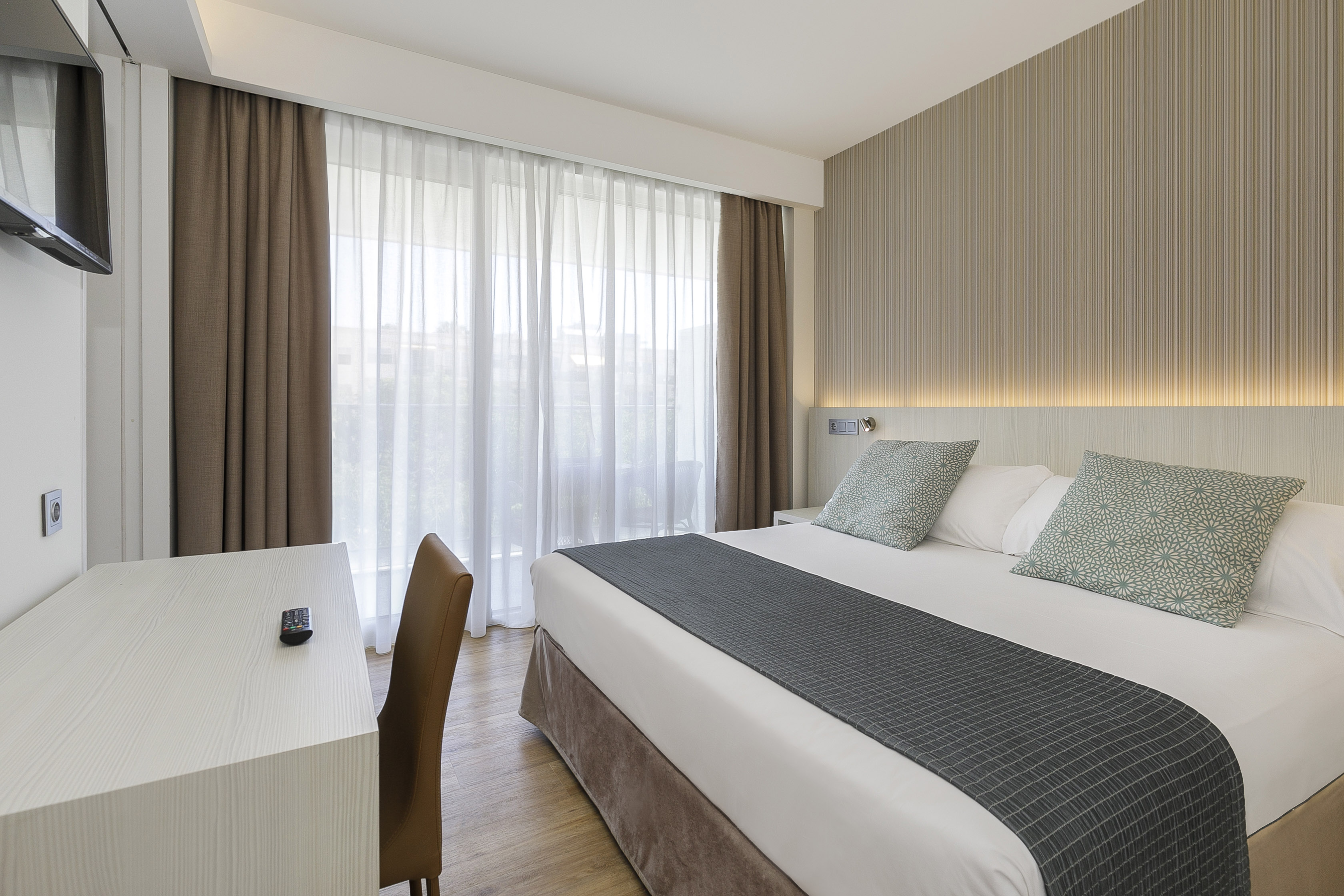 Modern hotel bedroom with large bed, desk, TV, and window with sheer curtains.