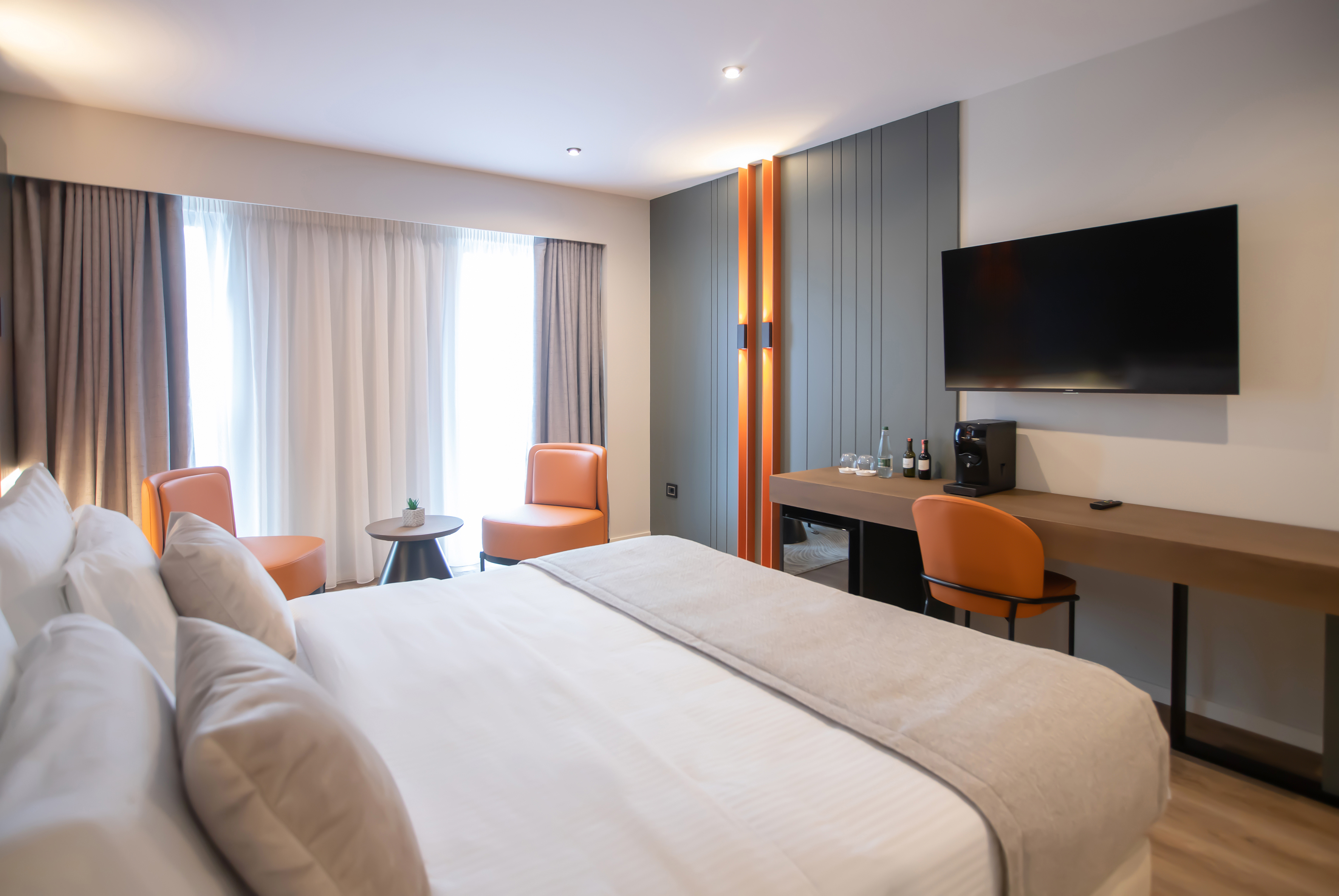 Modern hotel room with large bed, desk, TV, coffee maker, and orange accent chairs.