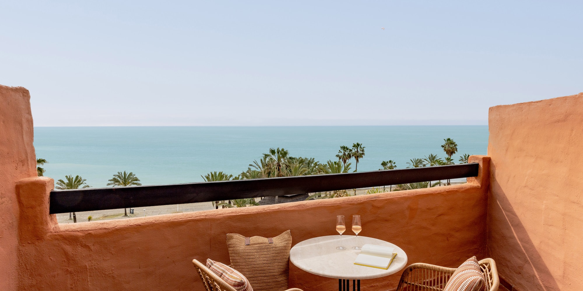 Terrace with sea view, palm trees, wicker chairs, and table with drinks.