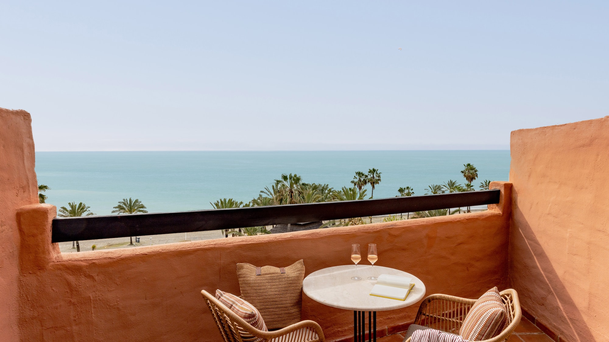 Terrace with sea view, palm trees, wicker chairs, and table with drinks.