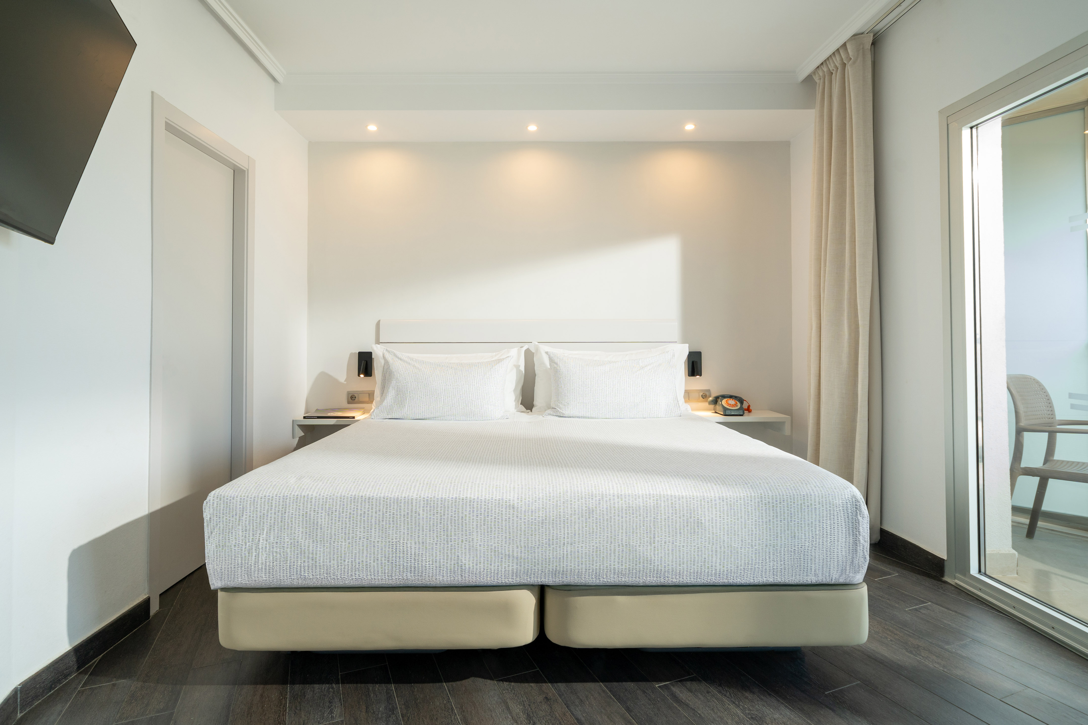 Modern hotel bedroom with white bed, bright lighting, and balcony access.