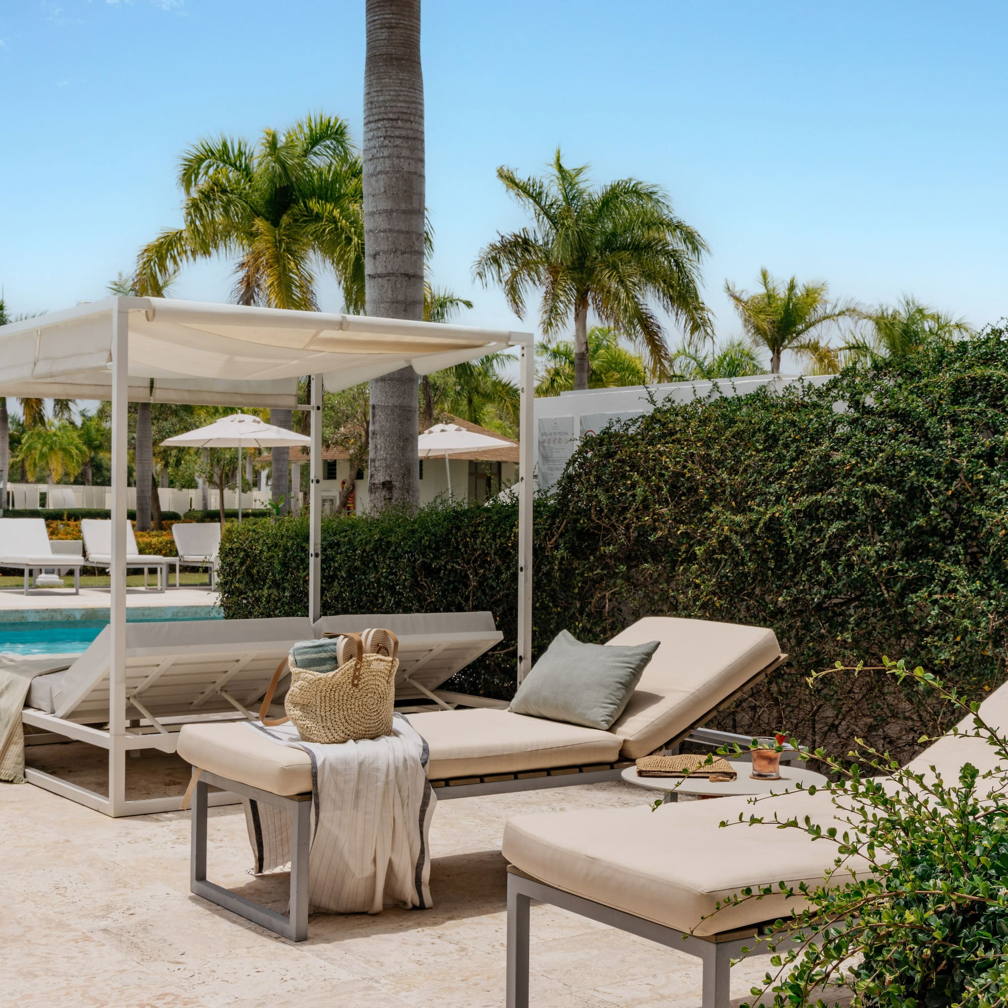 Sunny tropical resort pool with white daybeds, lounge chairs, and palm trees.