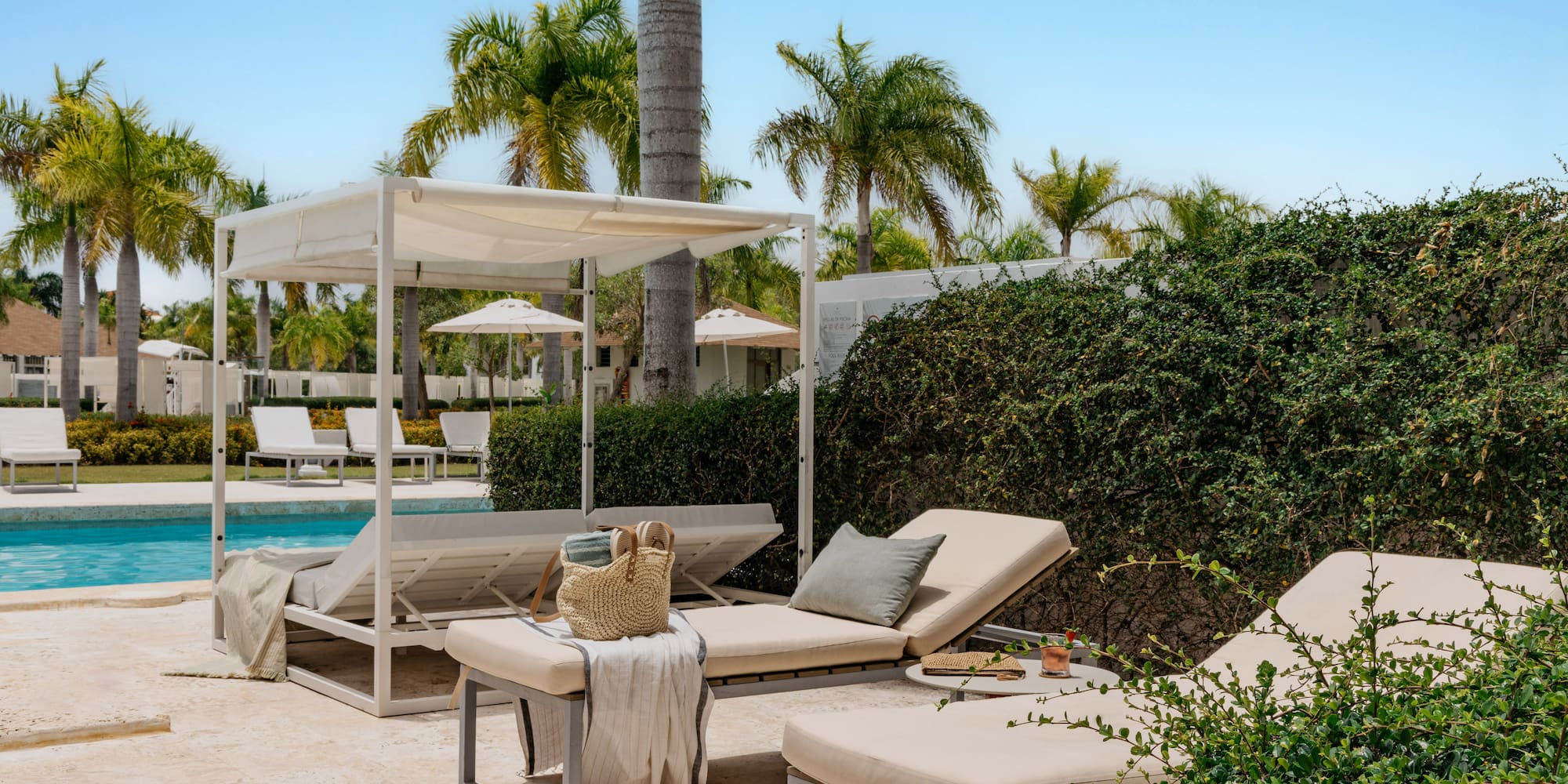 Sunny tropical resort pool with white daybeds, lounge chairs, and palm trees.