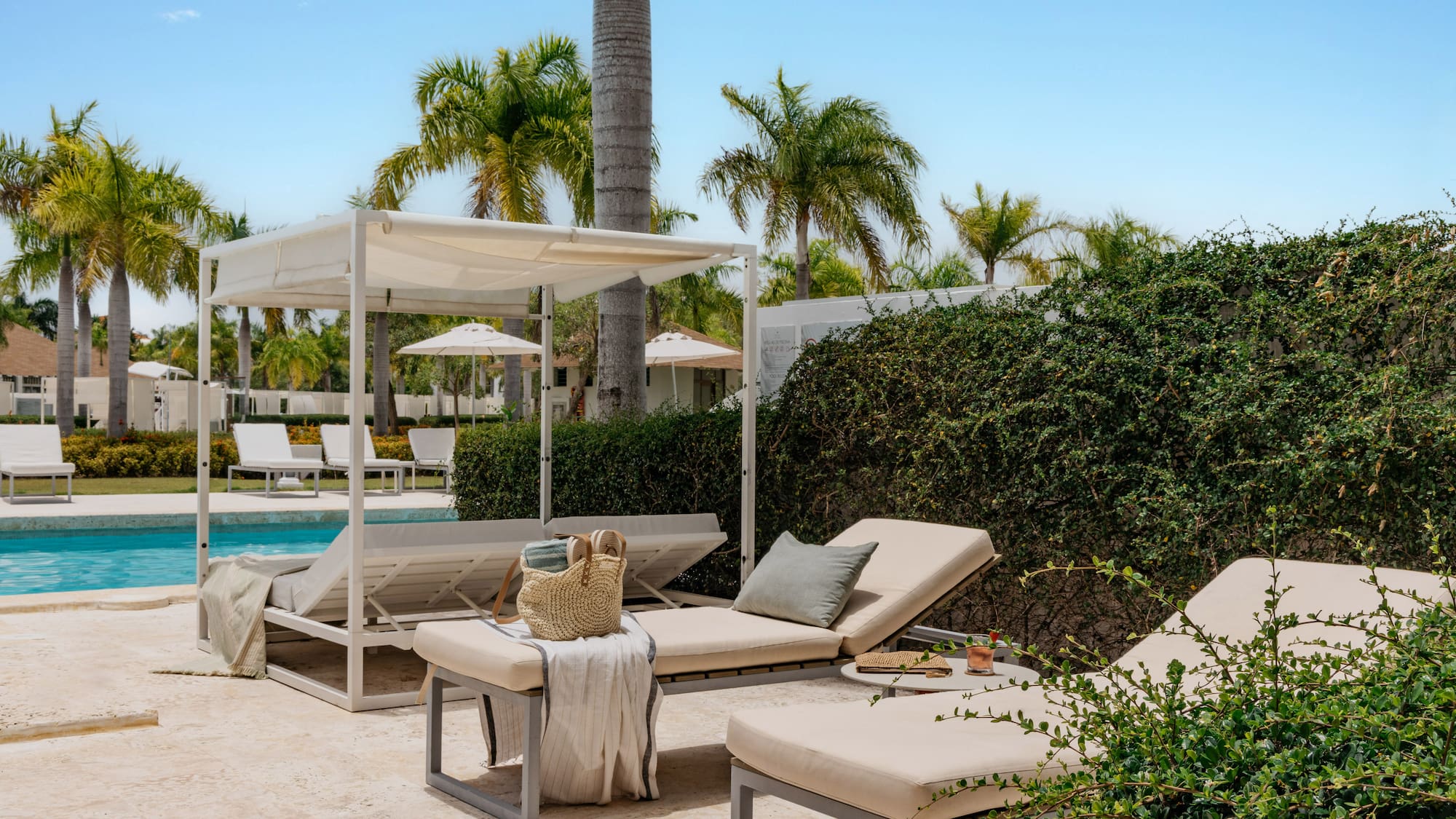 Sunny tropical resort pool with white daybeds, lounge chairs, and palm trees.