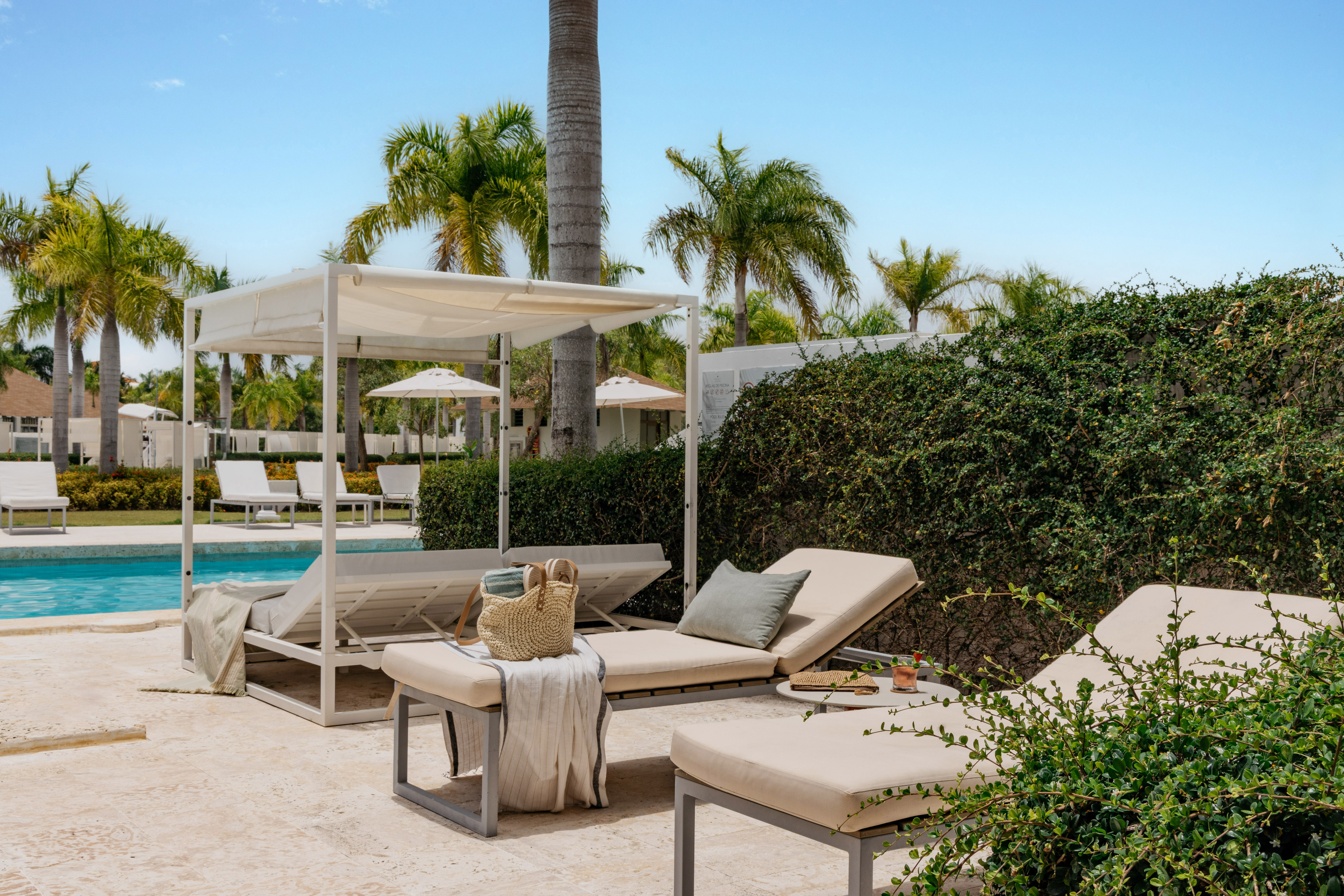 Sunny tropical resort pool with white daybeds, lounge chairs, and palm trees.