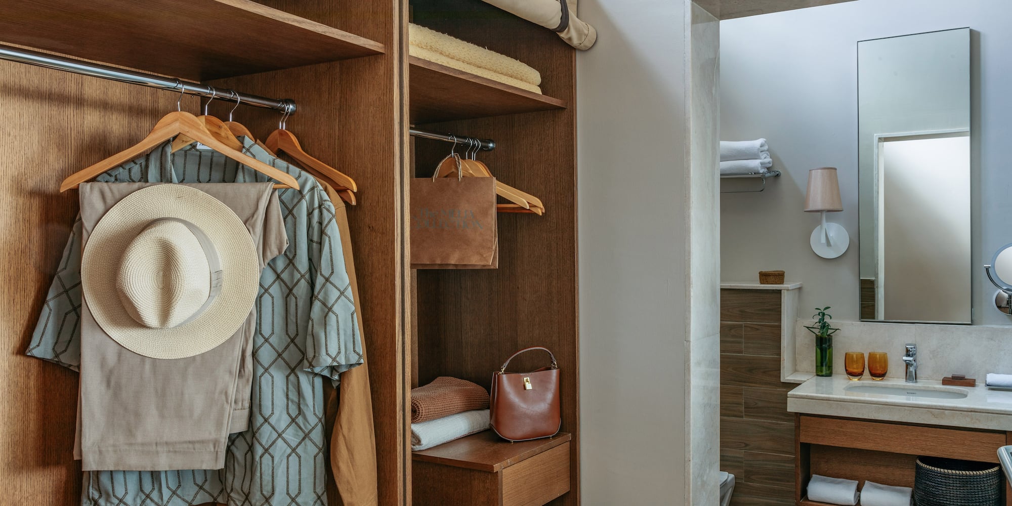 Hotel room closet with hanging clothes, straw hat, and bathroom vanity.
