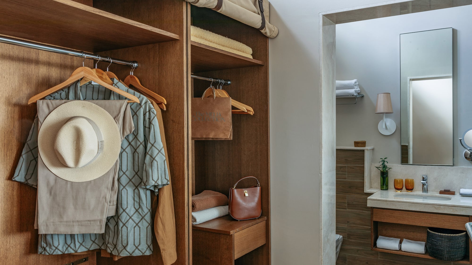 Hotel room closet with hanging clothes, straw hat, and bathroom vanity.