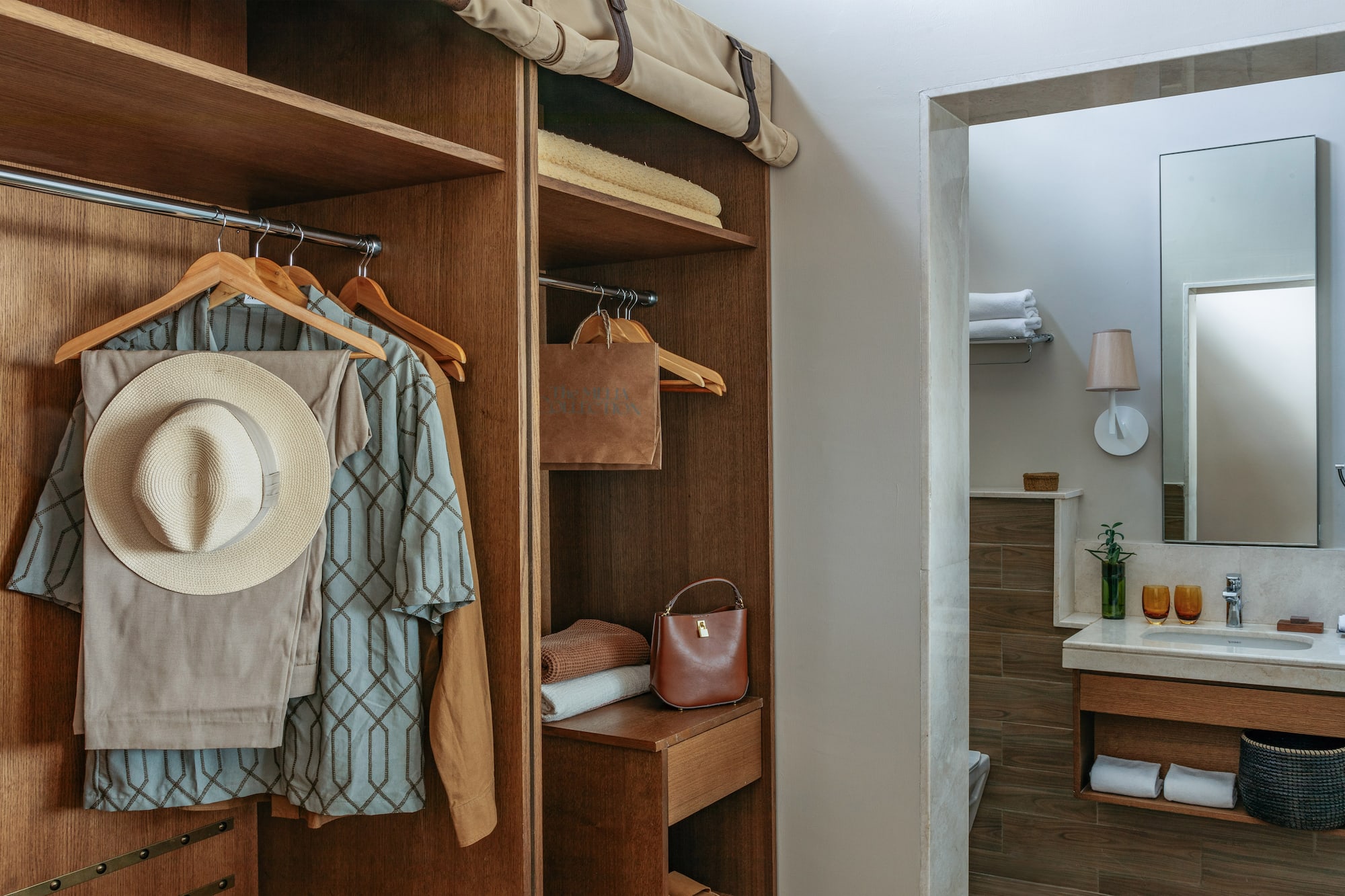Hotel room closet with hanging clothes, straw hat, and bathroom vanity.
