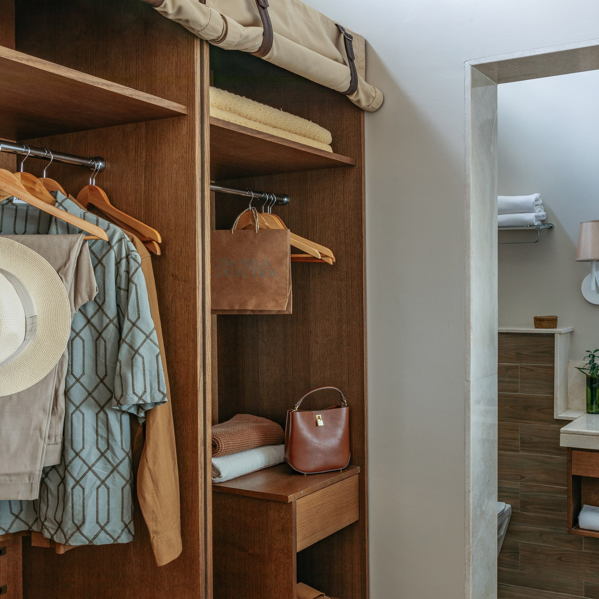 Hotel room closet with hanging clothes, straw hat, and bathroom vanity.