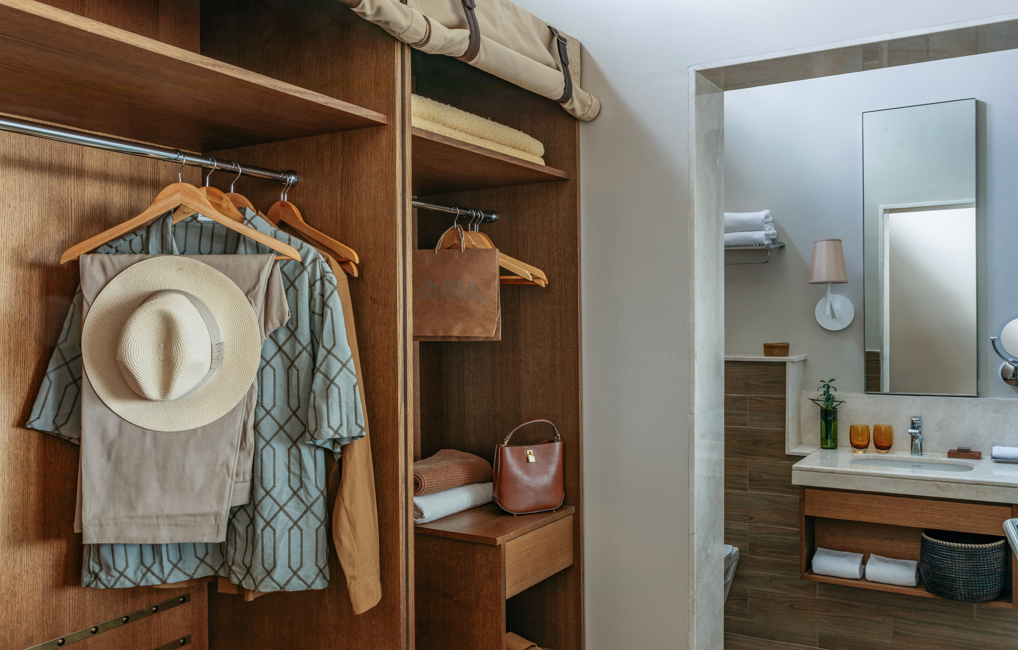 Hotel room closet with hanging clothes, straw hat, and bathroom vanity.