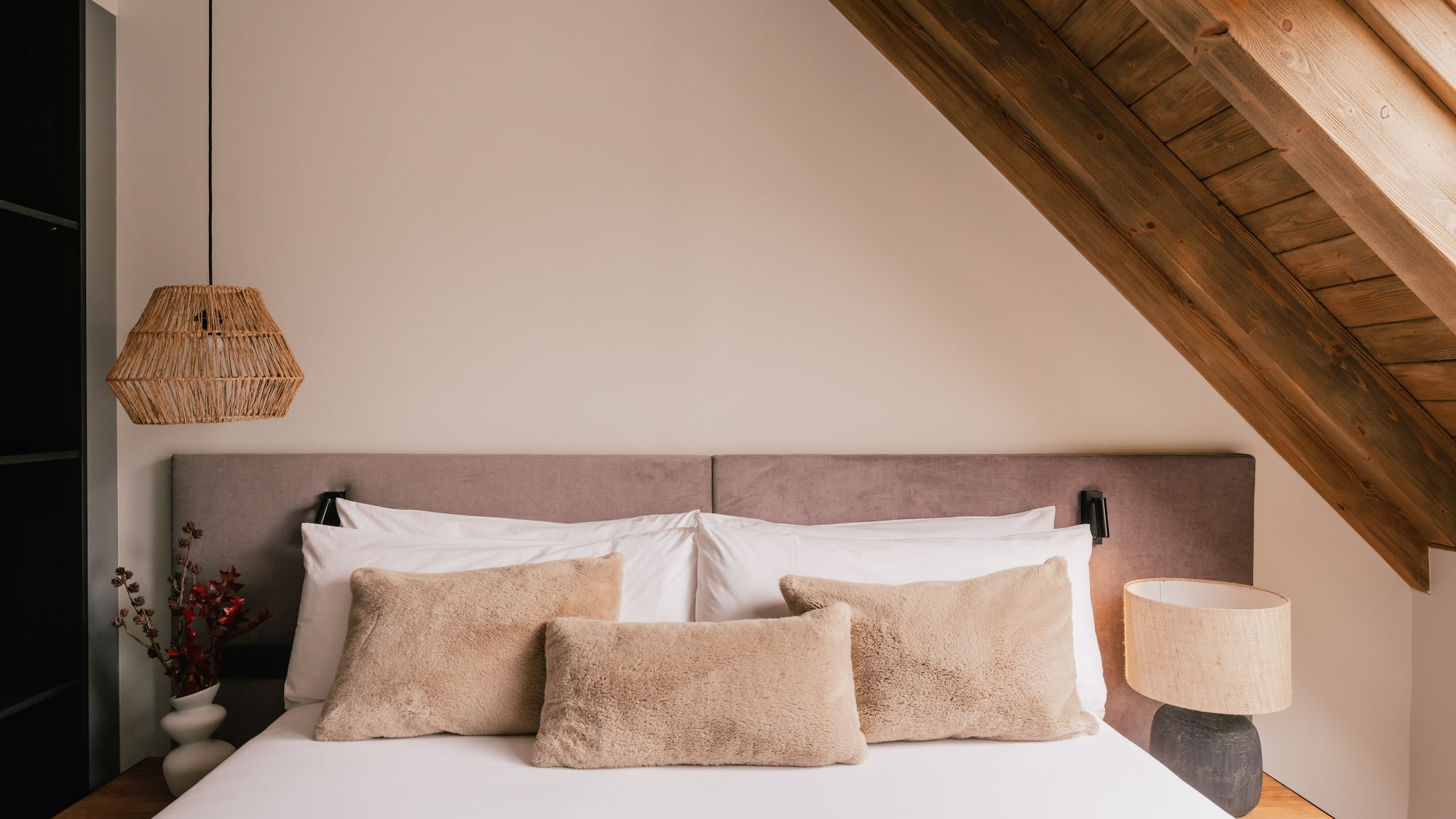 Cozy attic bedroom with wooden beams, fluffy pillows, and woven lamps.
