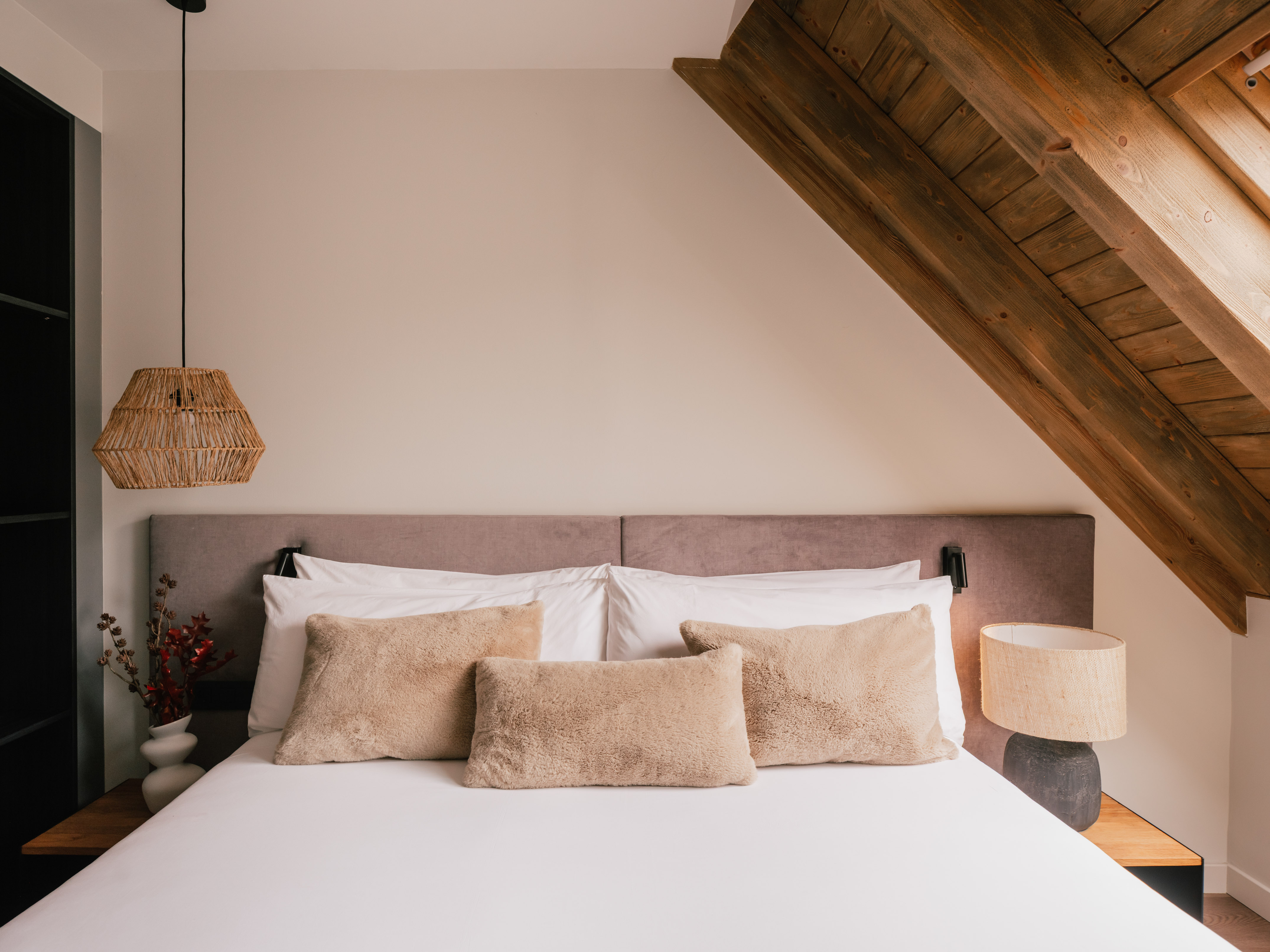 Cozy attic bedroom with wooden beams, fluffy pillows, and woven lamps.