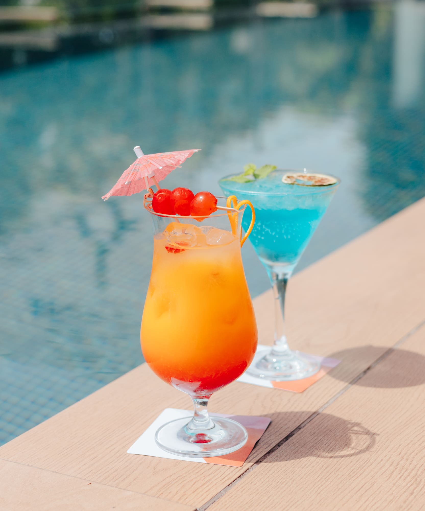 Orange and blue tropical cocktails with garnishes by a swimming pool.