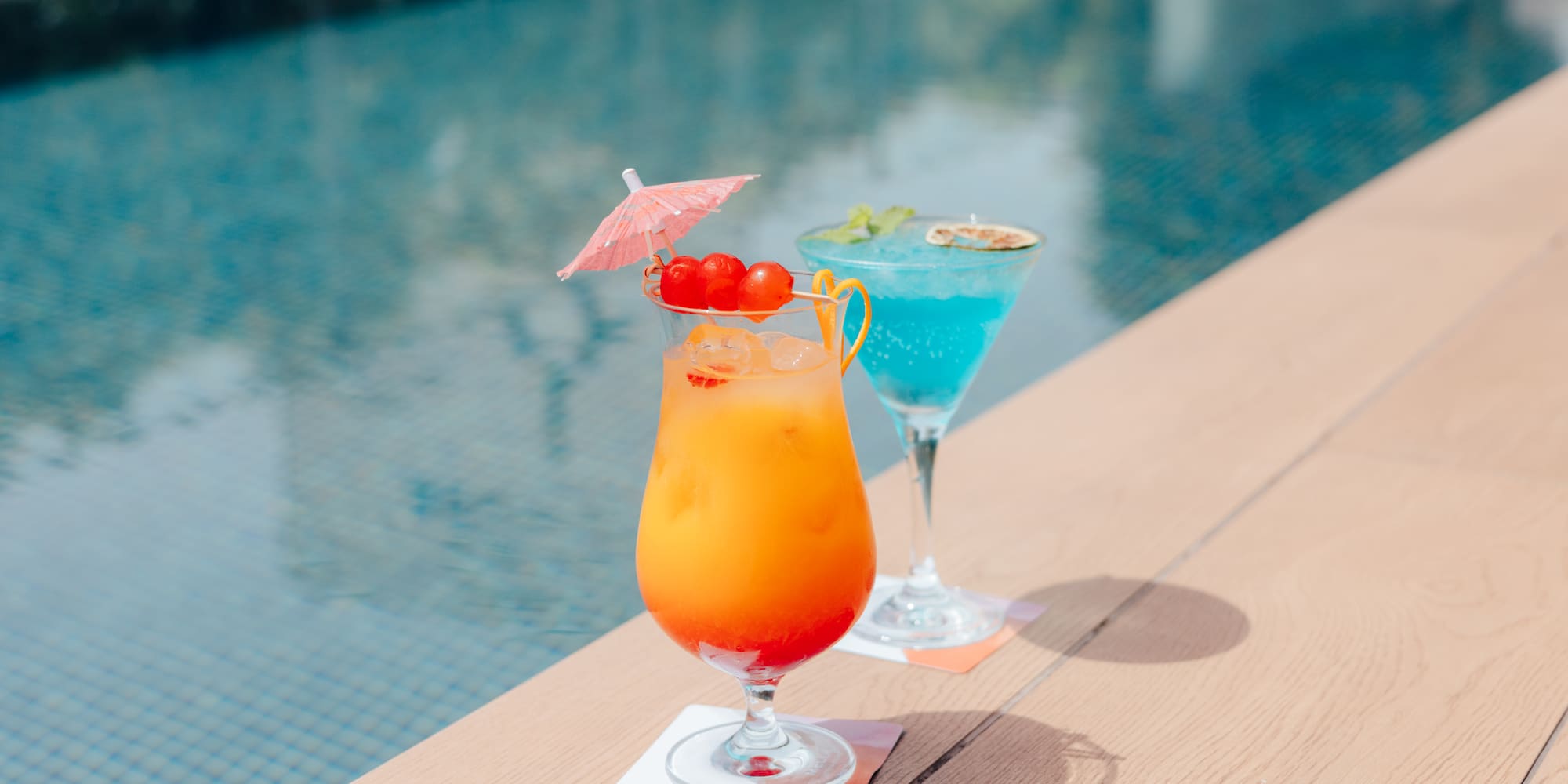 Orange and blue tropical cocktails with garnishes by a swimming pool.