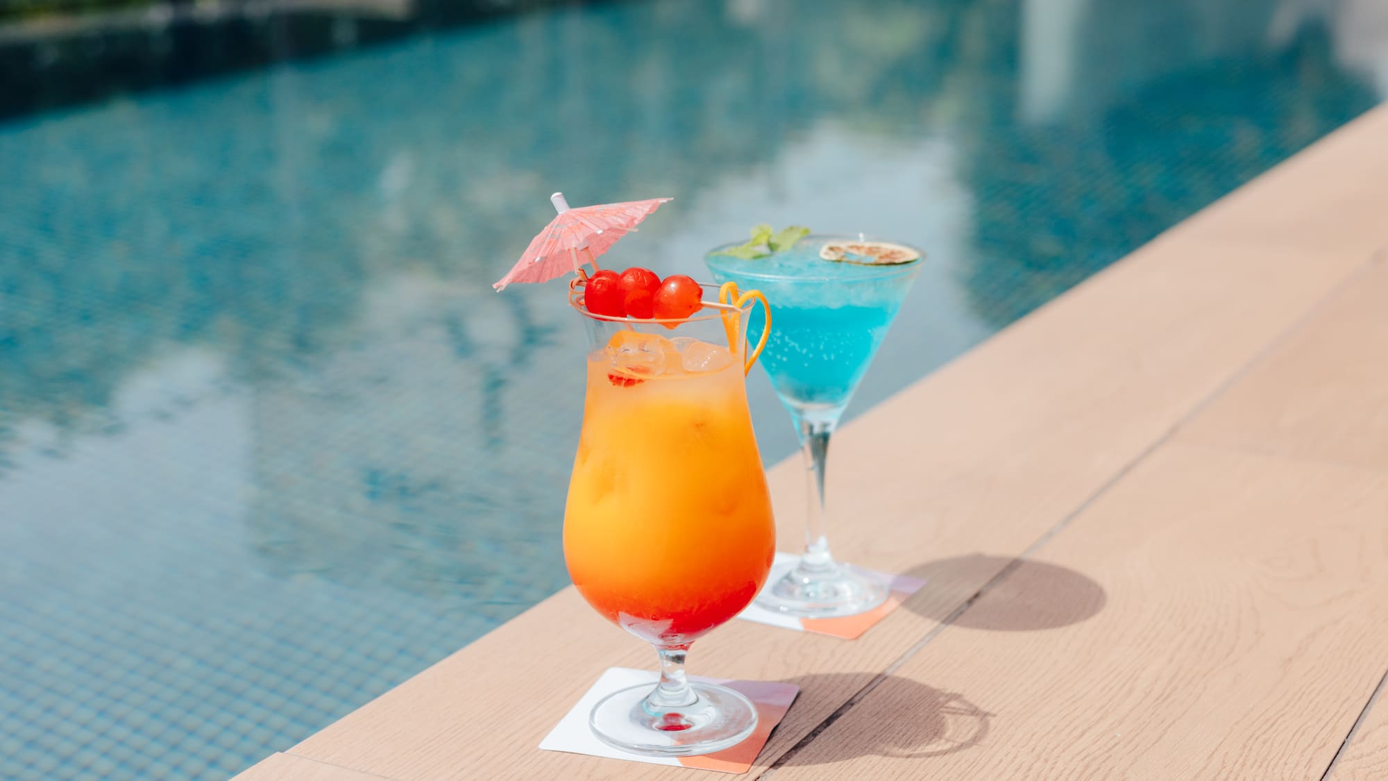 Two colorful tropical cocktails with garnishes by a bright blue pool.